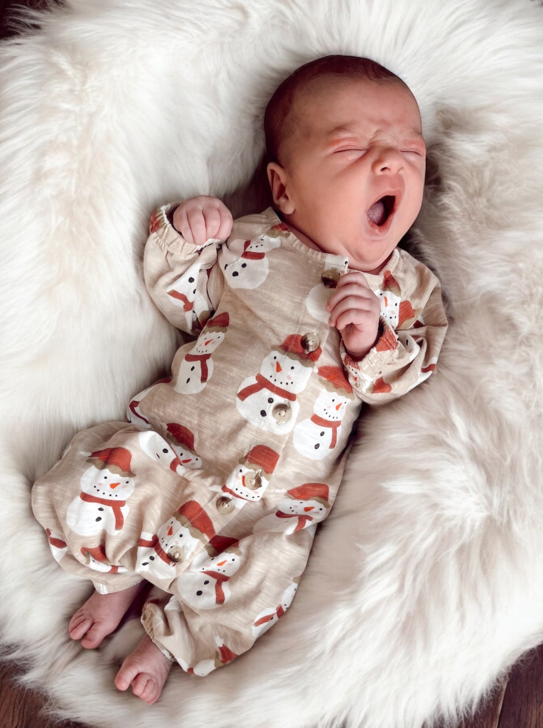 Baby in a snowman-patterned outfit yawning on a fluffy white rug.