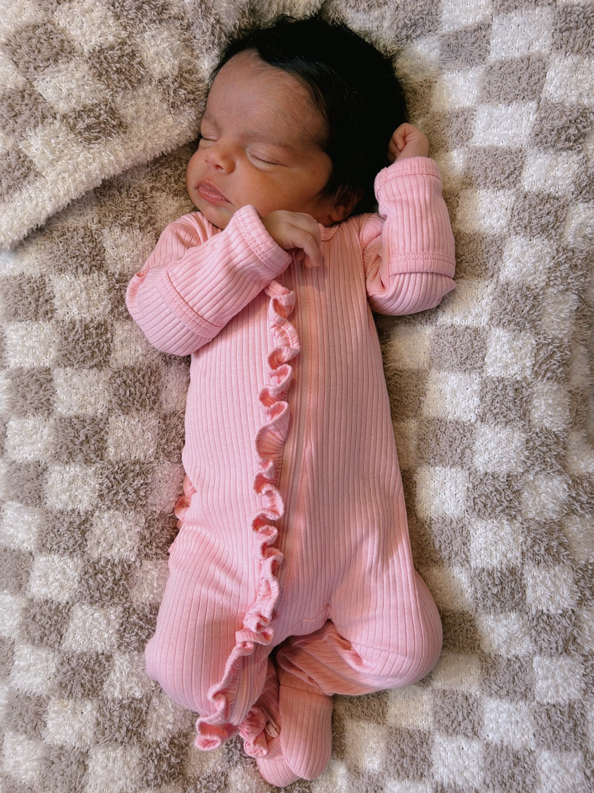 Newborn baby peacefully sleeping in a pink ribbed onesie on a soft checkered blanket.