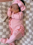 Sleeping infant in pink onesie with ruffles and matching bow, resting on a cozy, textured blanket.