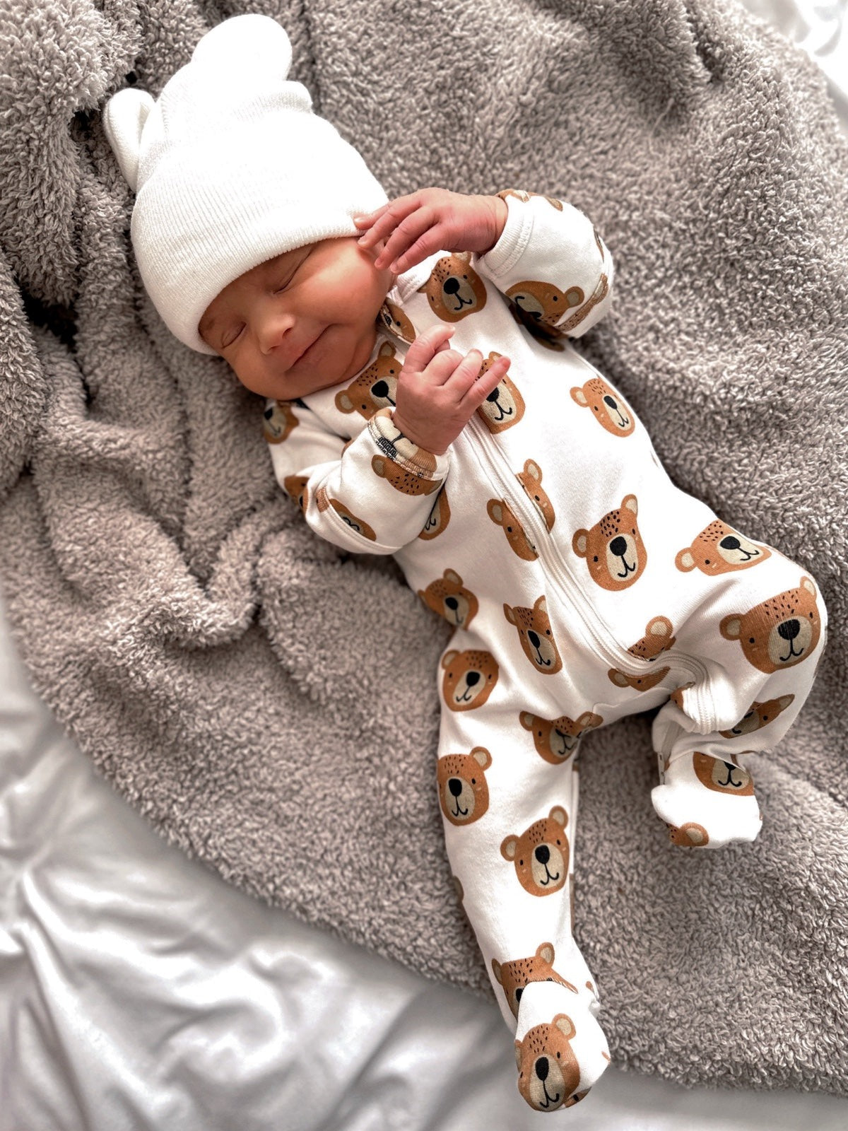 Smiling baby in bear-patterned pajamas and a white hat, resting on a soft gray blanket.