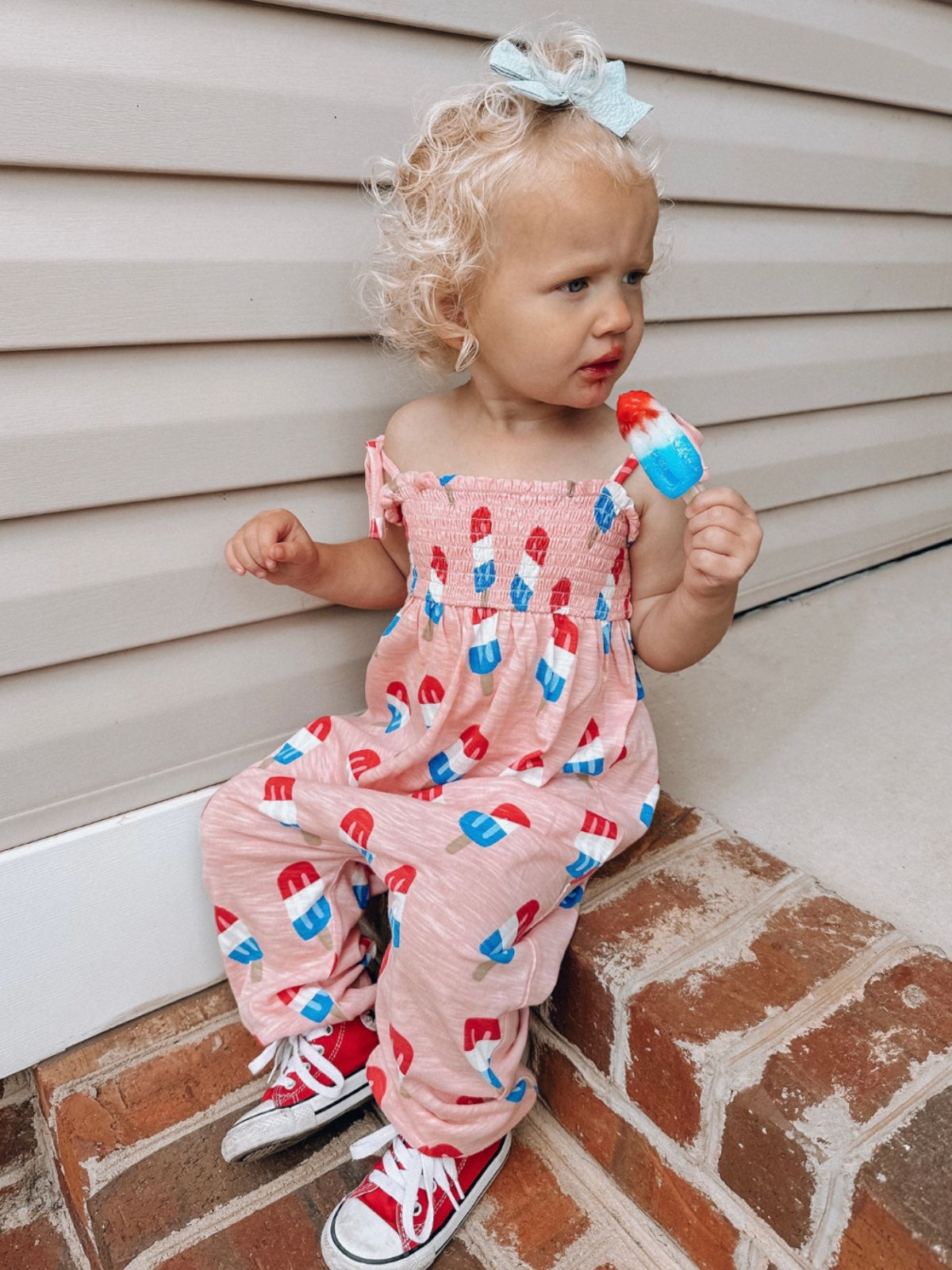 Toddler sitting on bricks wearing a pink popsicle-patterned outfit, holding a colorful popsicle.