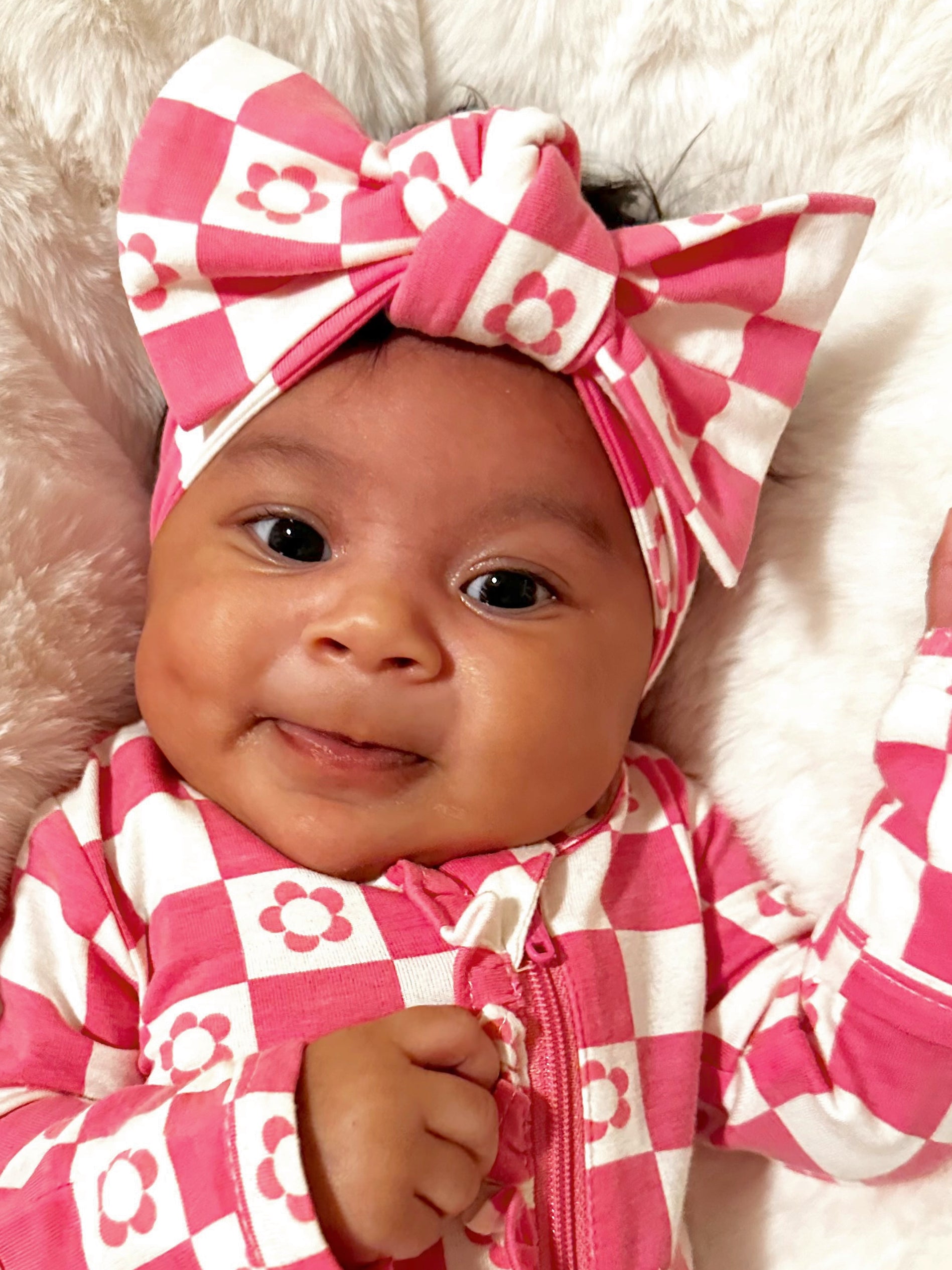 Smiling baby wearing a pink checkered outfit and a large bow, resting on a fluffy white surface.
