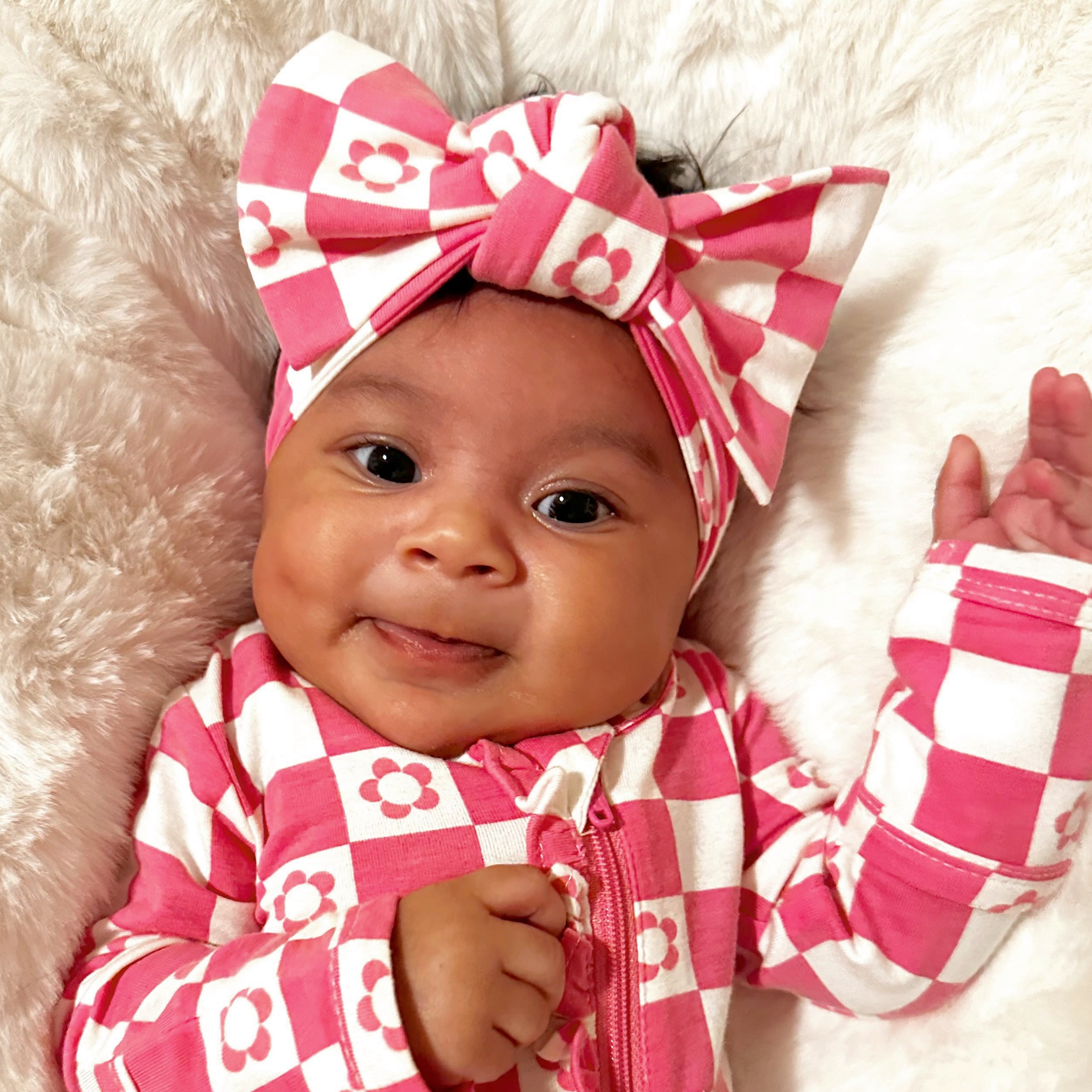 Smiling baby wearing a pink checkered outfit and a large bow, resting on a fluffy white surface.