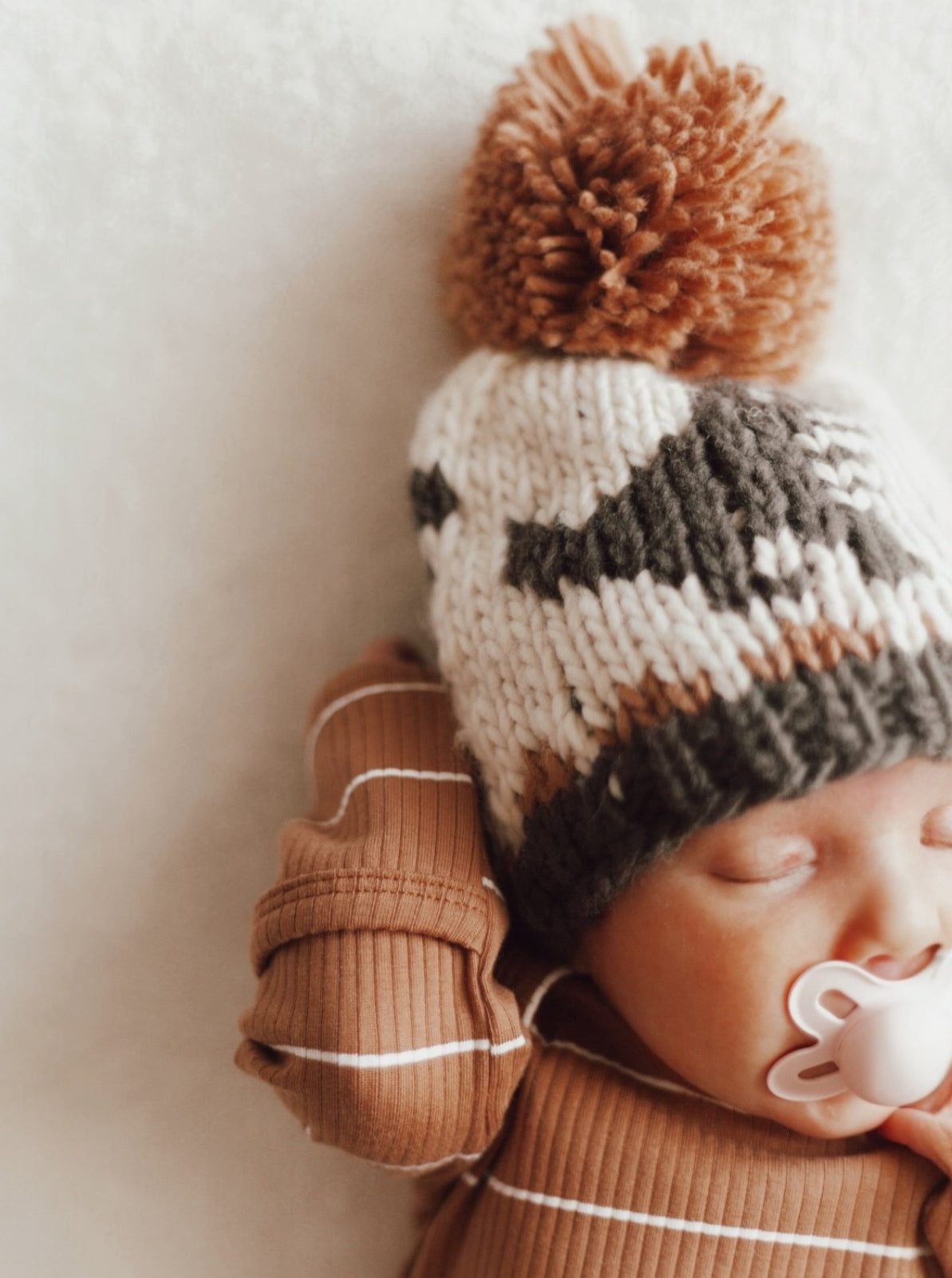 Baby wearing a knitted hat with pom-pom, peacefully sleeping with a pacifier, on a soft, light backdrop.