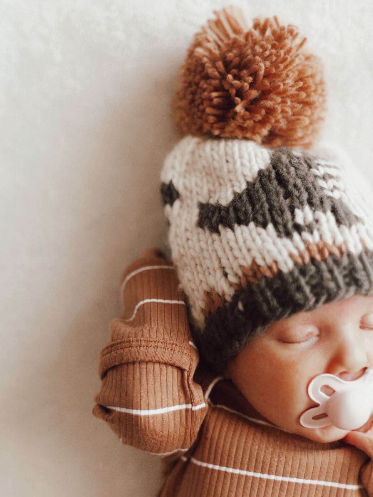 Baby wearing a knitted hat with pom-pom, peacefully sleeping with a pacifier, on a soft, light backdrop.