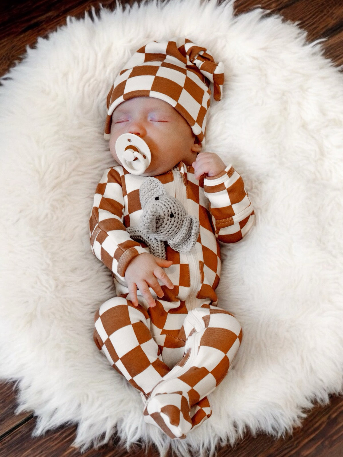 Sleeping baby in brown and white checkered outfit, cuddling a toy, resting on a fluffy white blanket.