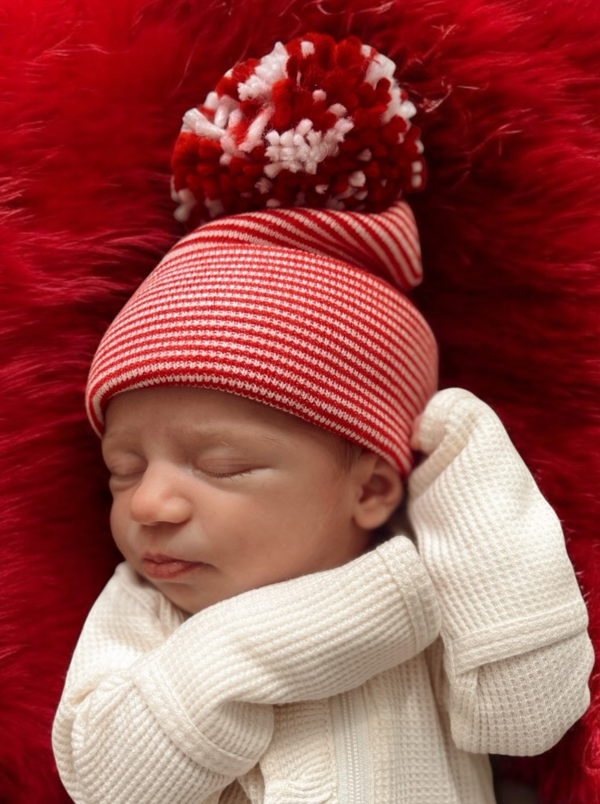 Newborn baby girl sleeping on a soft red background, wearing a red and white striped hat with a pom-pom.
