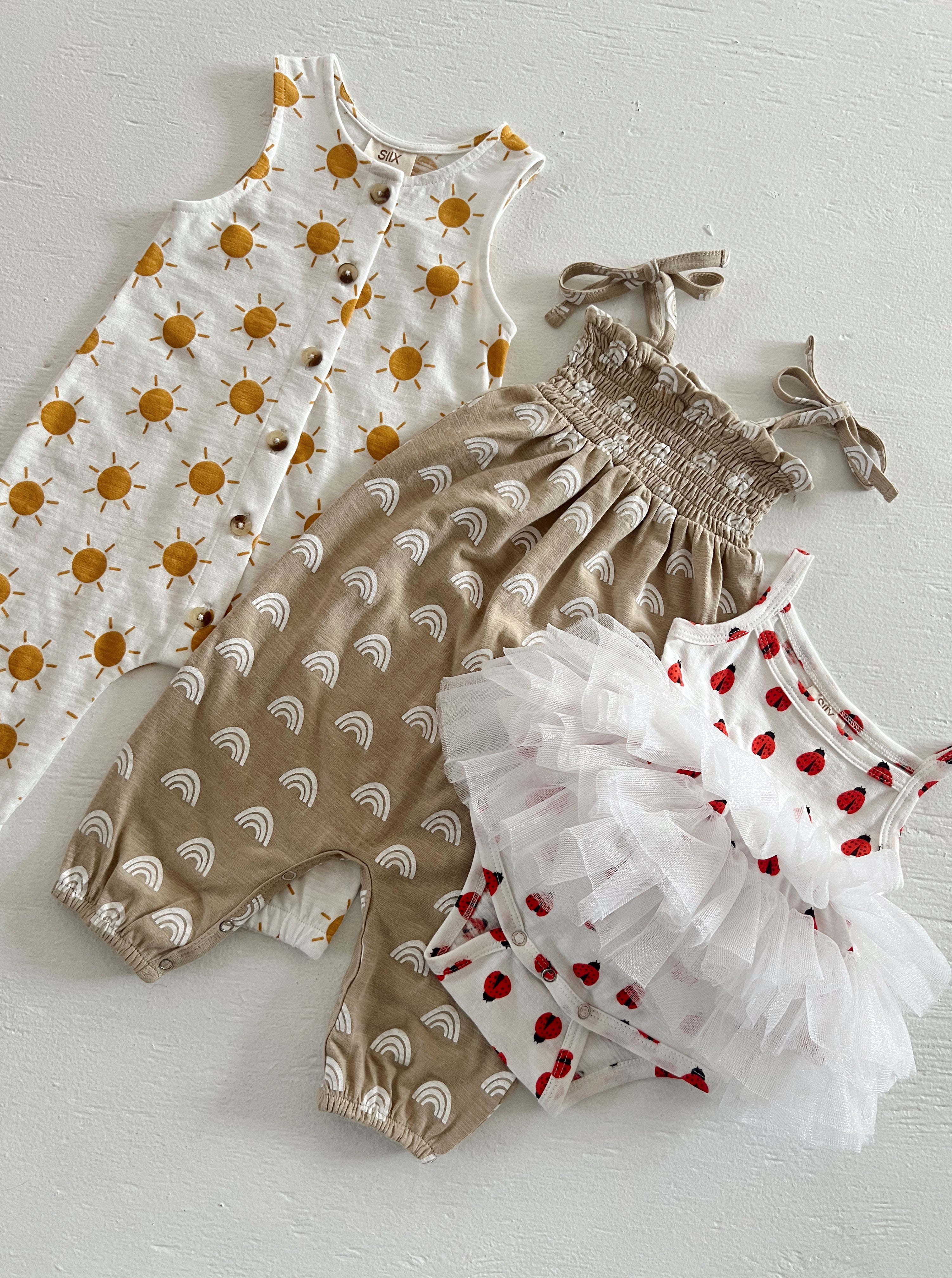 Three children's outfits: a sun-patterned dress, rainbow romper, and a tulle ladybug tank top.