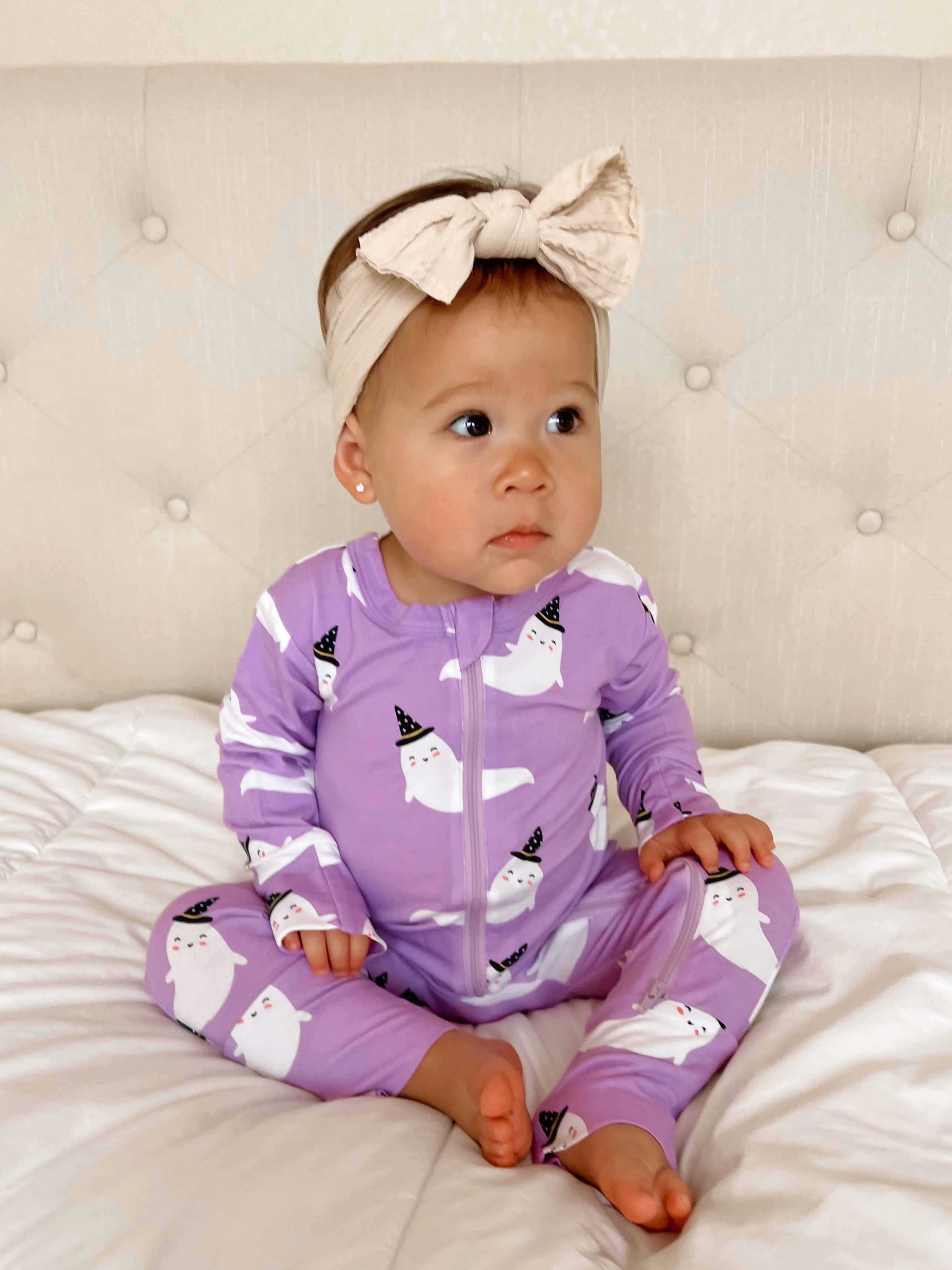 Toddler in a purple ghost-patterned onesie with a large bow headband, sitting on a white quilted bedspread.