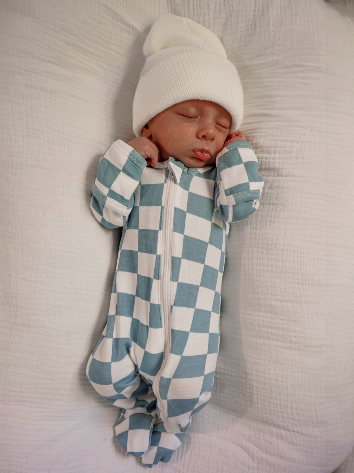 Sleeping baby in a checkered onesie and white beanie on a soft blanket.