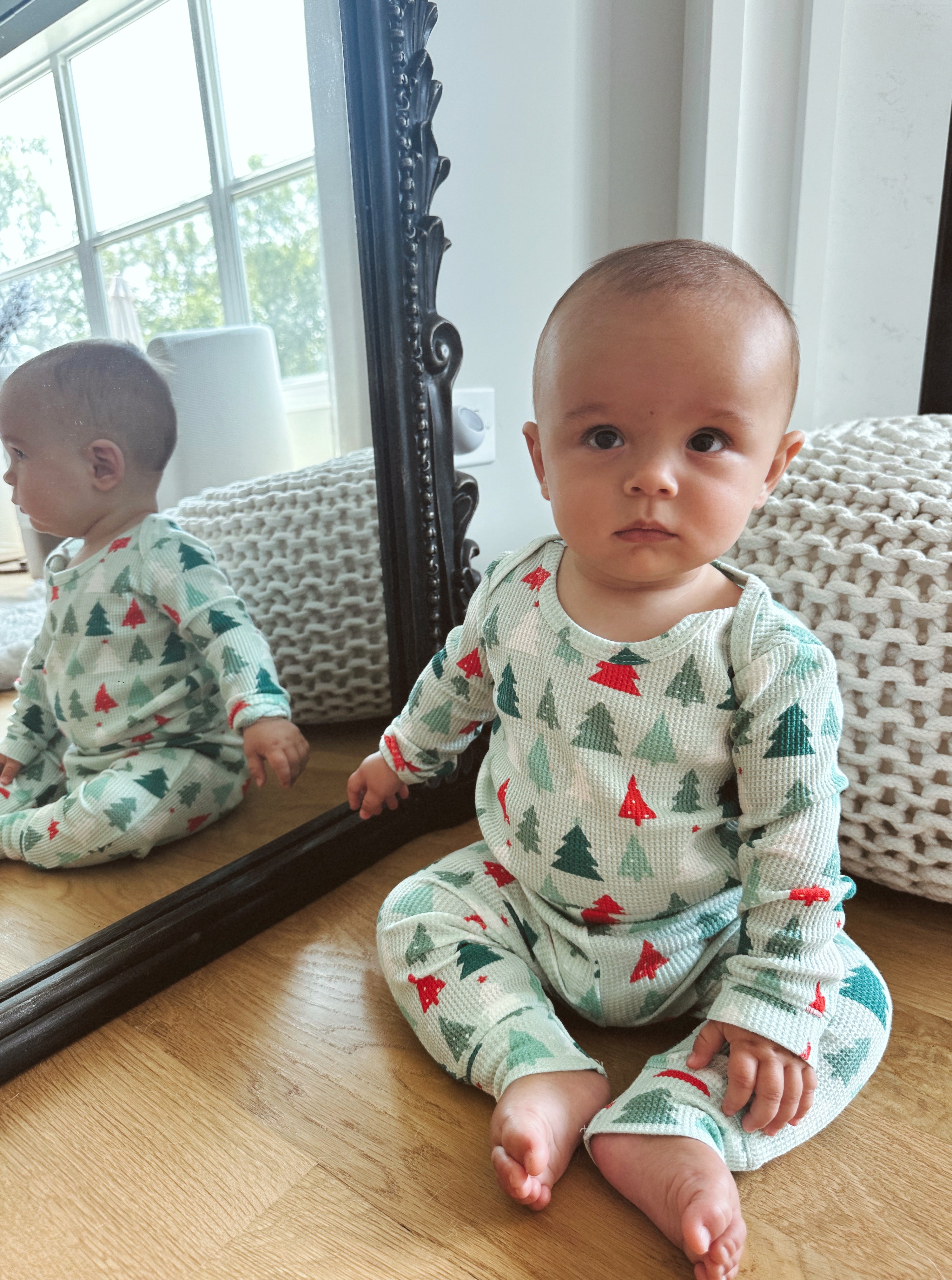 Baby in festive pajamas sits on the floor, reflecting in a mirror, with a cozy blanket nearby and natural light.