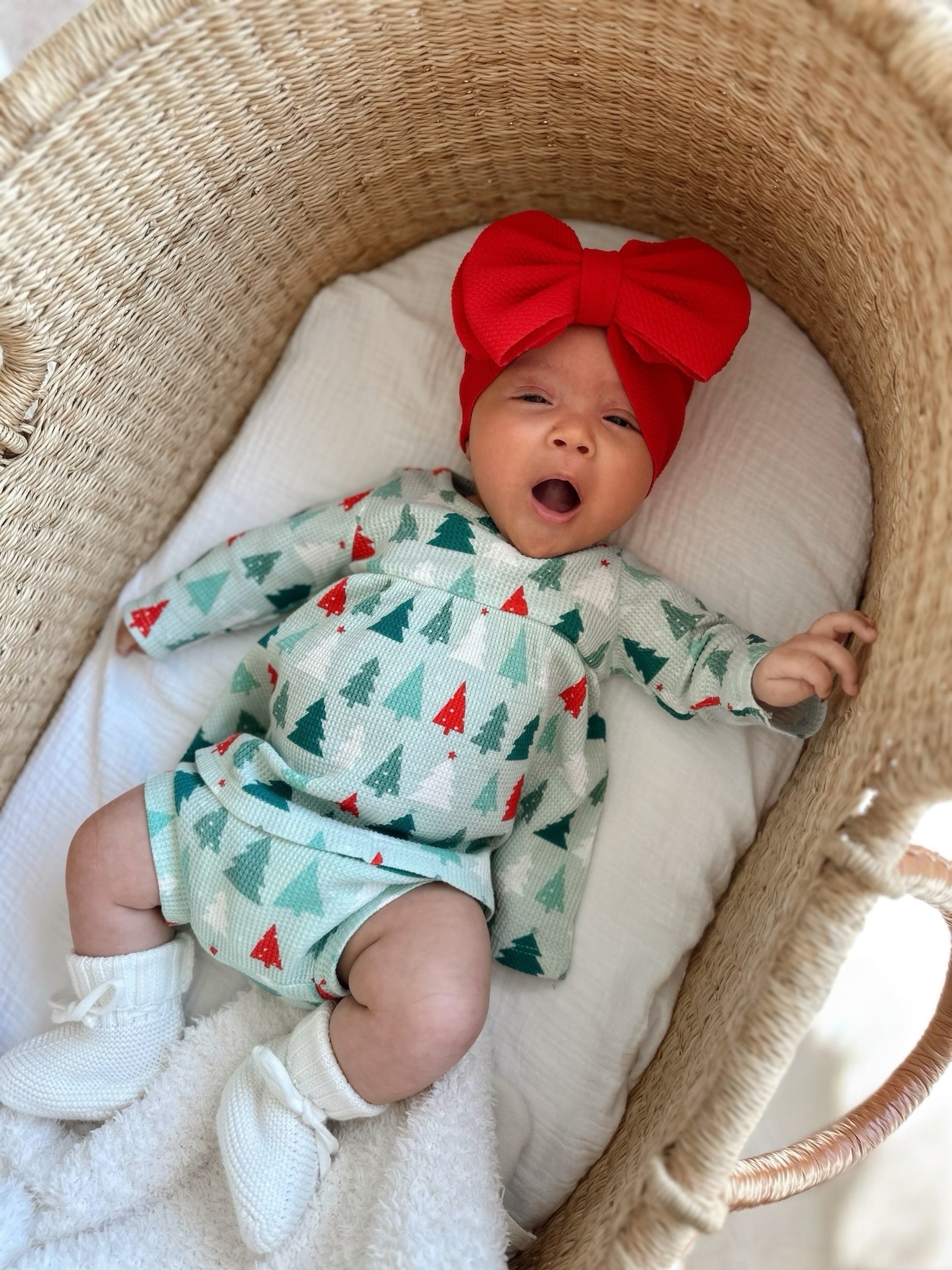Baby in a cozy bassinet, wearing a festive tree-patterned outfit and a bright red bow headband, yawning adorably.