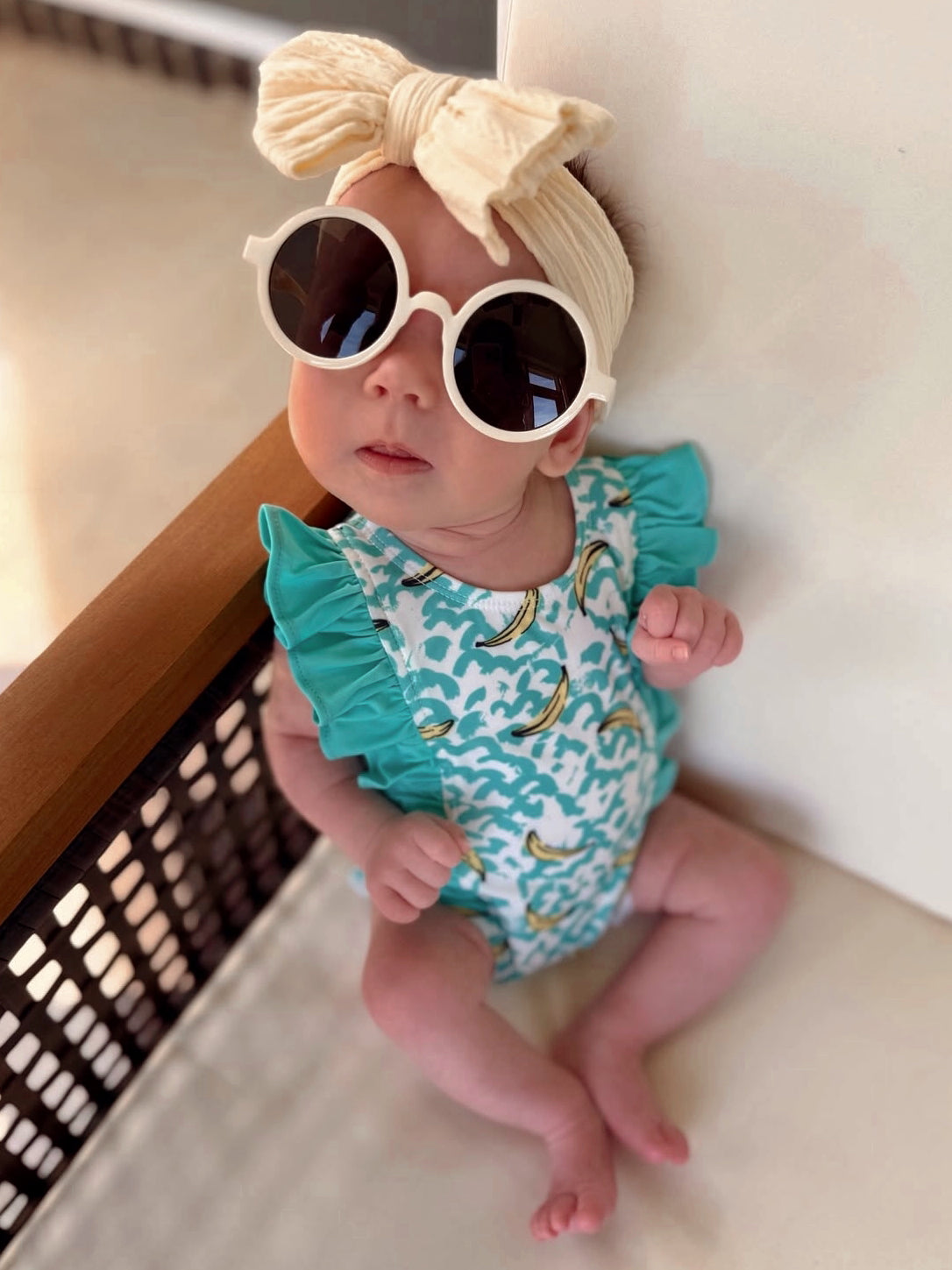 Baby wearing oversized sunglasses and a headband, sitting on a chair in a colorful swimsuit with a banana pattern.