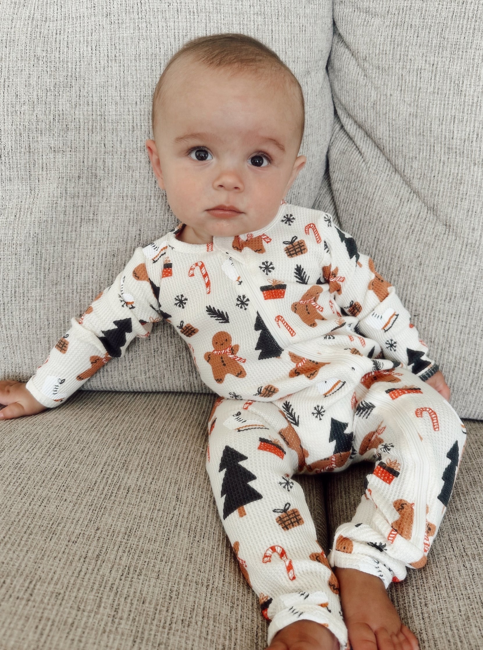 Baby sitting on a sofa, wearing festive pajamas with gingerbread and Christmas patterns.