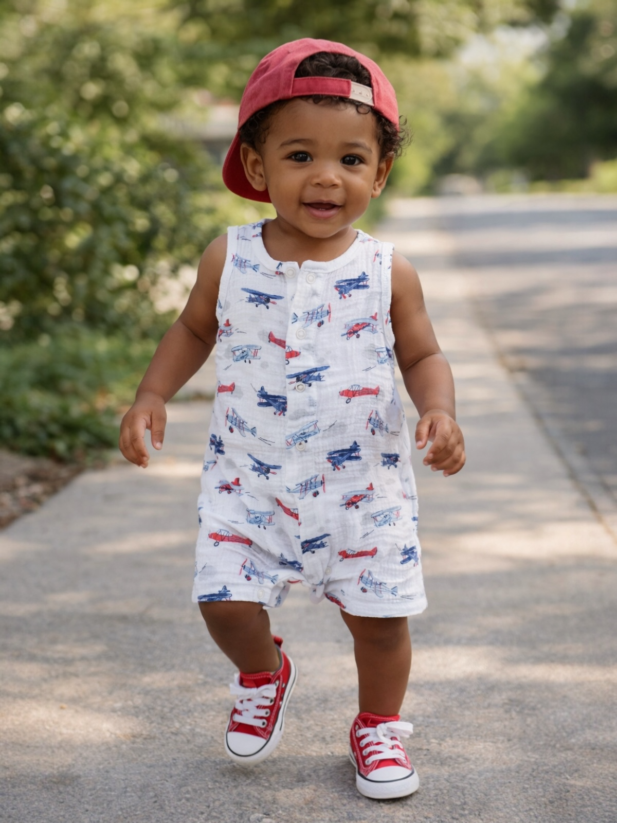 Smiling toddler in a colorful airplane-patterned romper, red sneakers, and a backward cap walking on a sunny pathway.