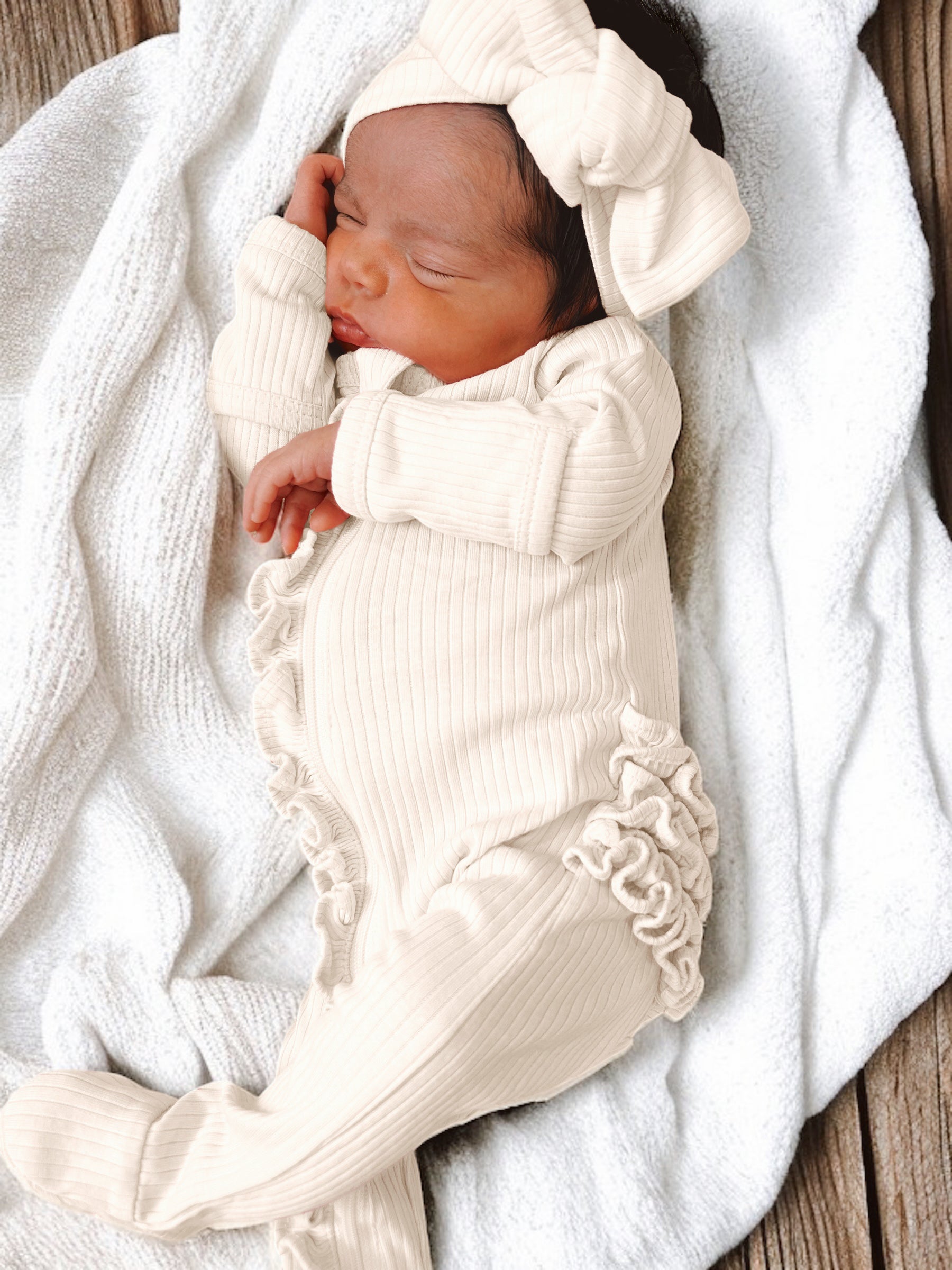 Newborn girl sleeping in a cream outfit and matching headband on a soft blanket.