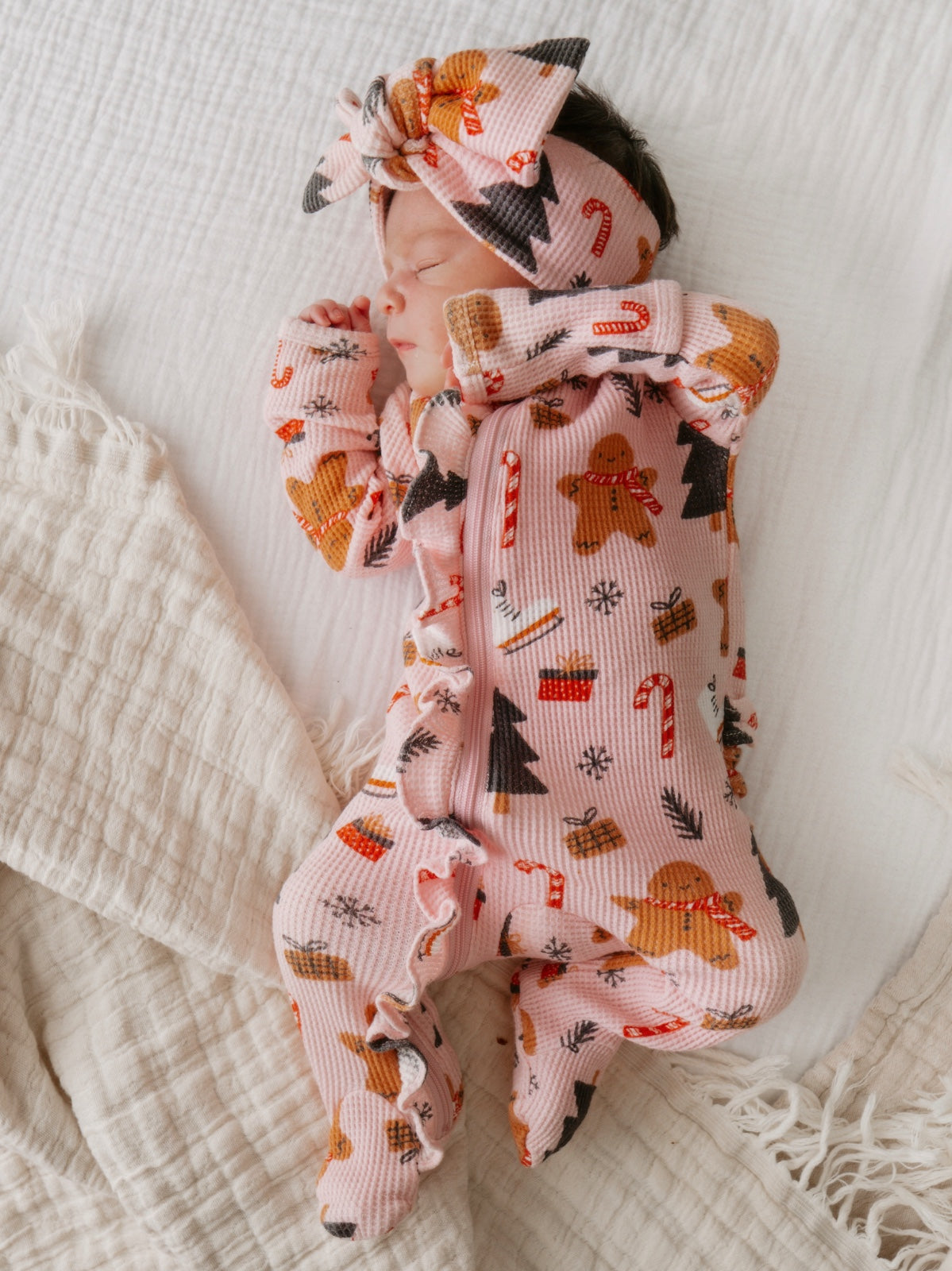 Sleeping baby in festive pink pajamas with gingerbread and candy cane patterns, lying on a soft white blanket.
