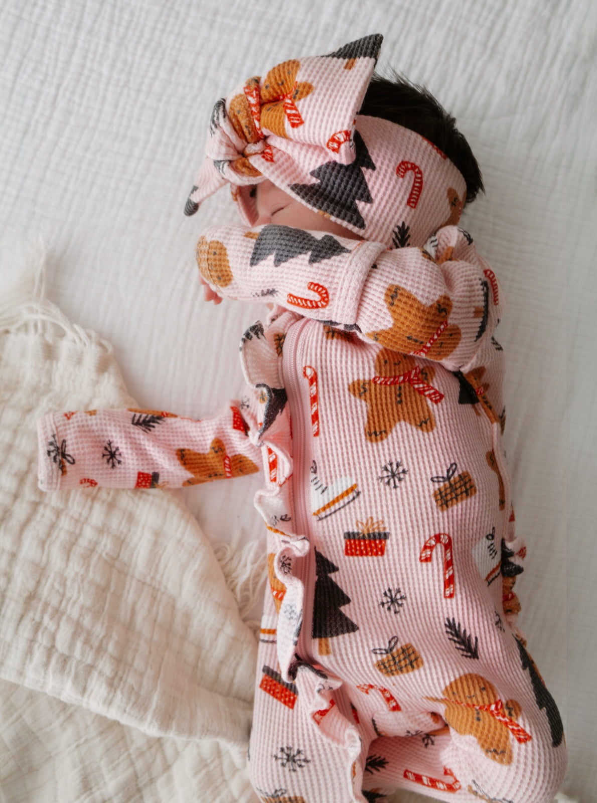 Baby in holiday-themed outfit with gingerbread and candy cane patterns, resting on a soft blanket.