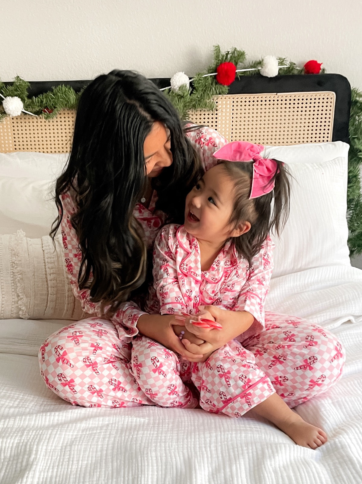 Mother and daughter share a joyful moment in matching pink pajamas, seated on a cozy bed with festive decor.