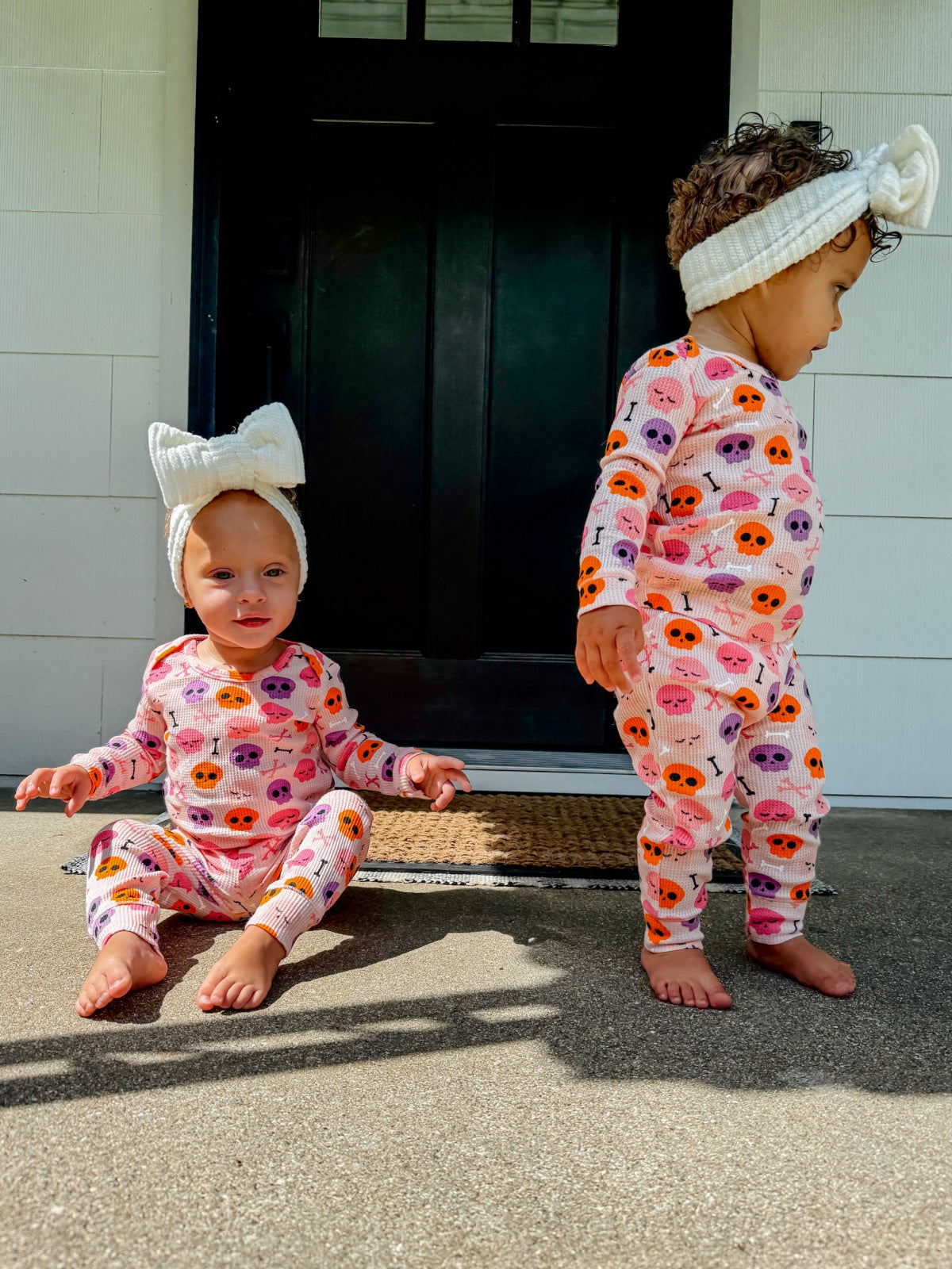 Two toddlers in colorful skull-patterned pajamas, one sitting and one standing near a black door.