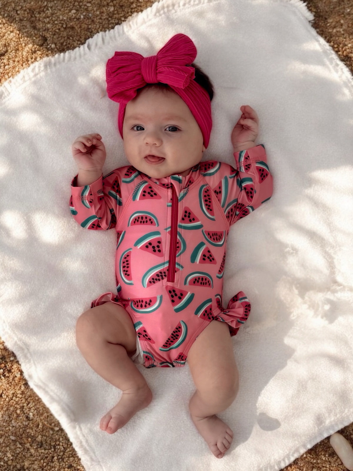 Baby with a pink watermelon-printed swimsuit and a matching headband, lying on a soft white blanket outdoors.