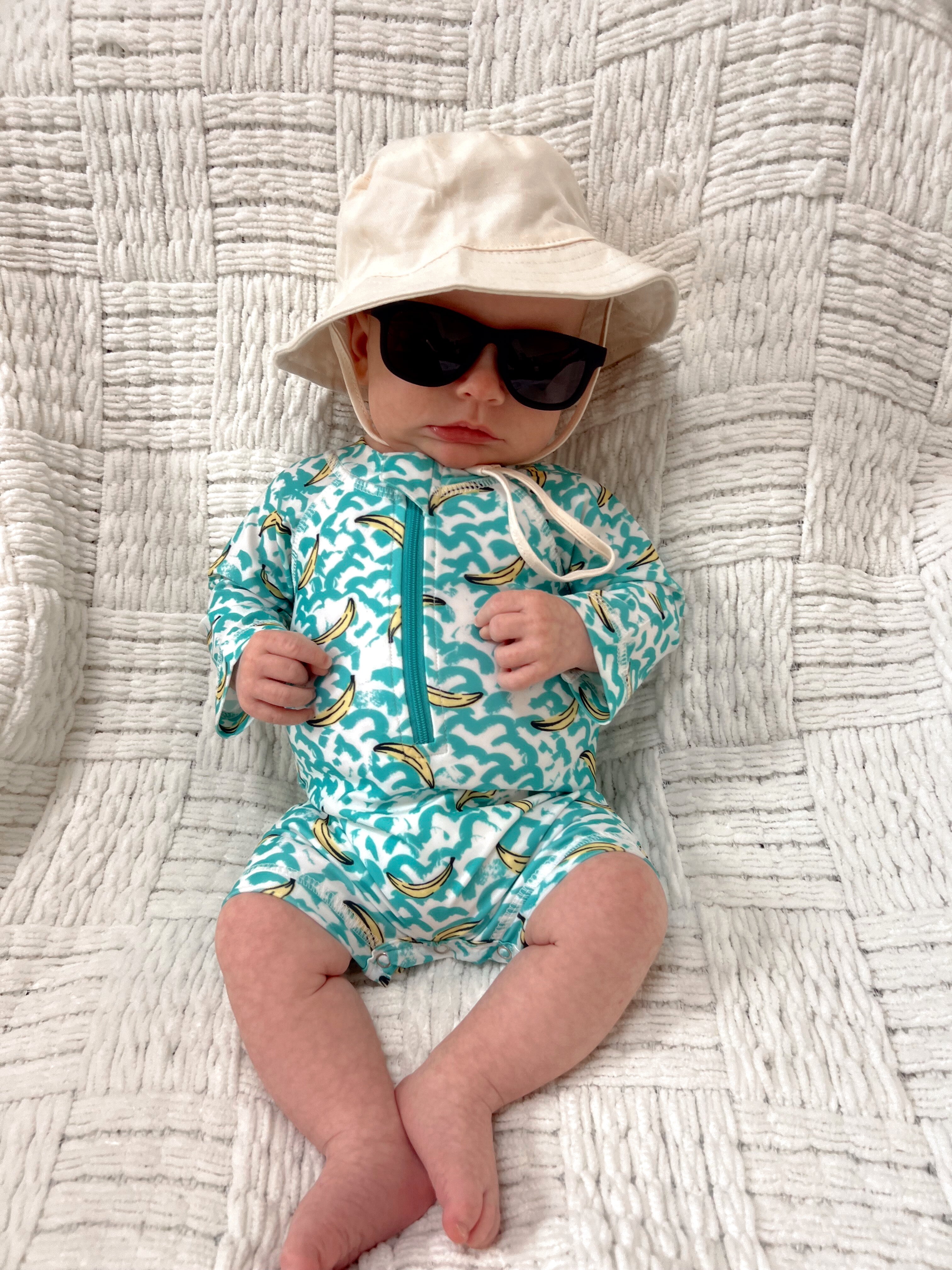 Baby in a colorful swimsuit, white bucket hat, and sunglasses, sitting on a textured blanket.