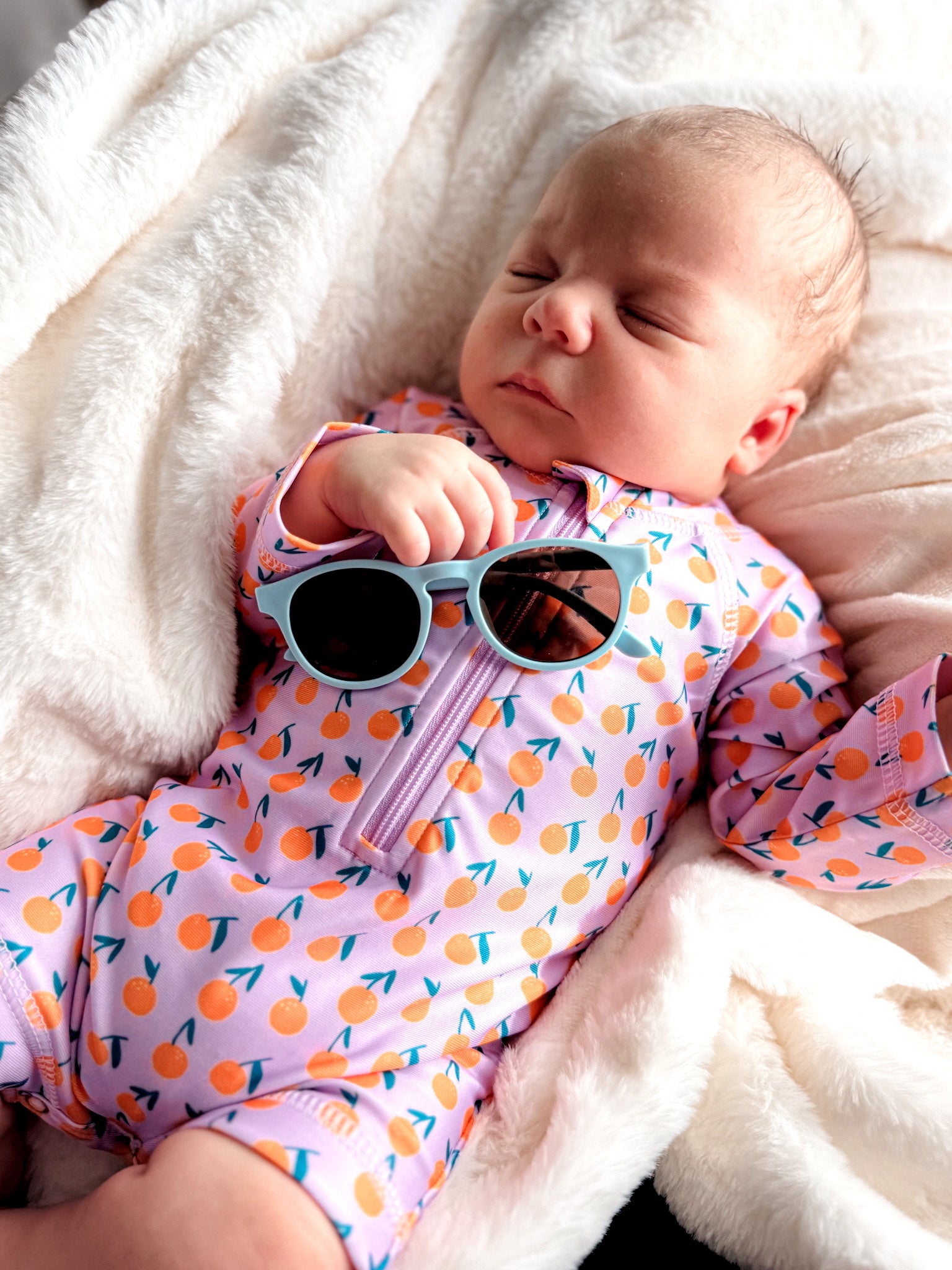 A sleeping baby in bright orange-patterned swimwear holds sunglasses while resting on a soft blanket.