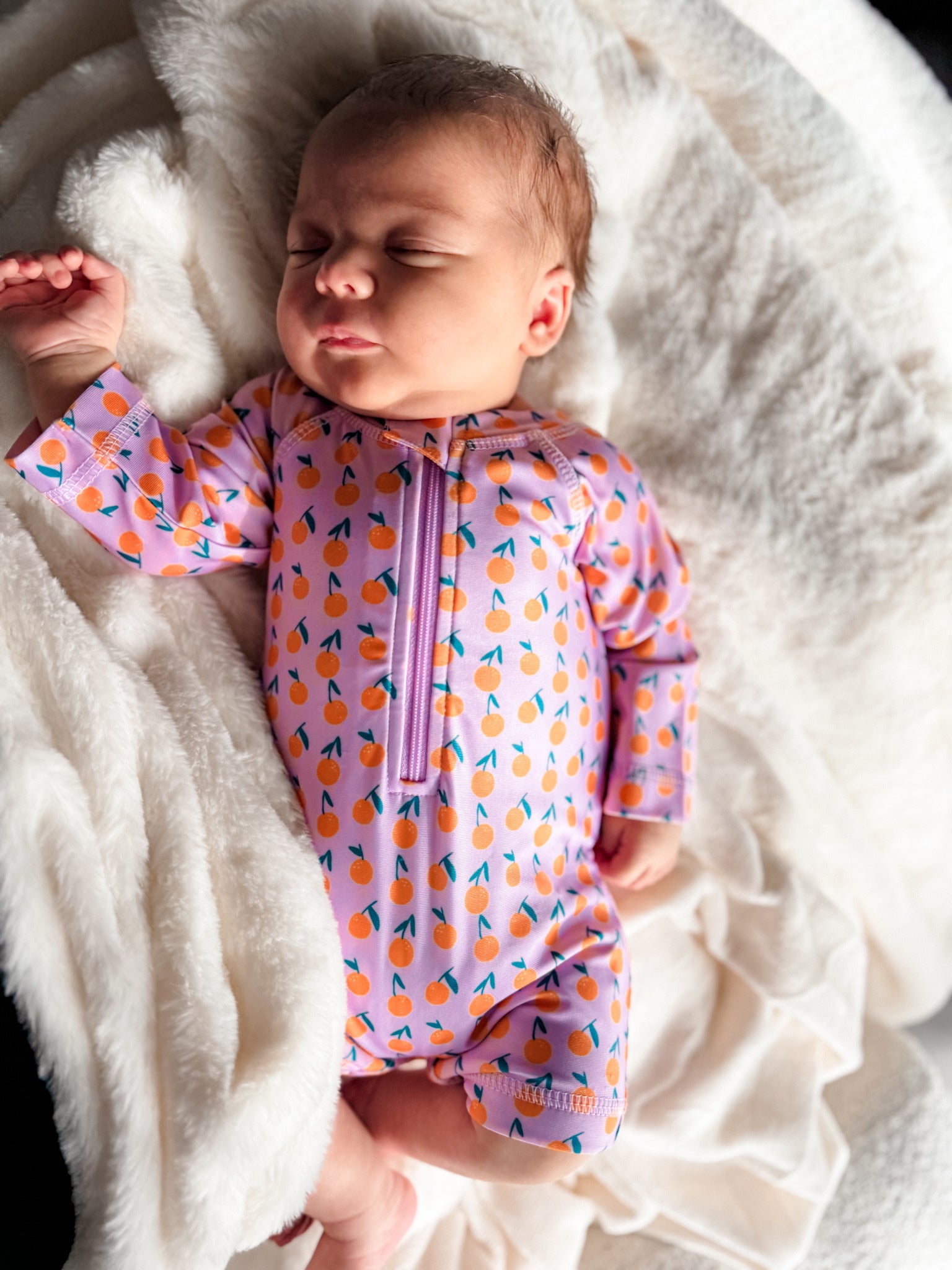 Sleeping baby in a colorful onesie with fruit patterns, resting peacefully on a soft white blanket.