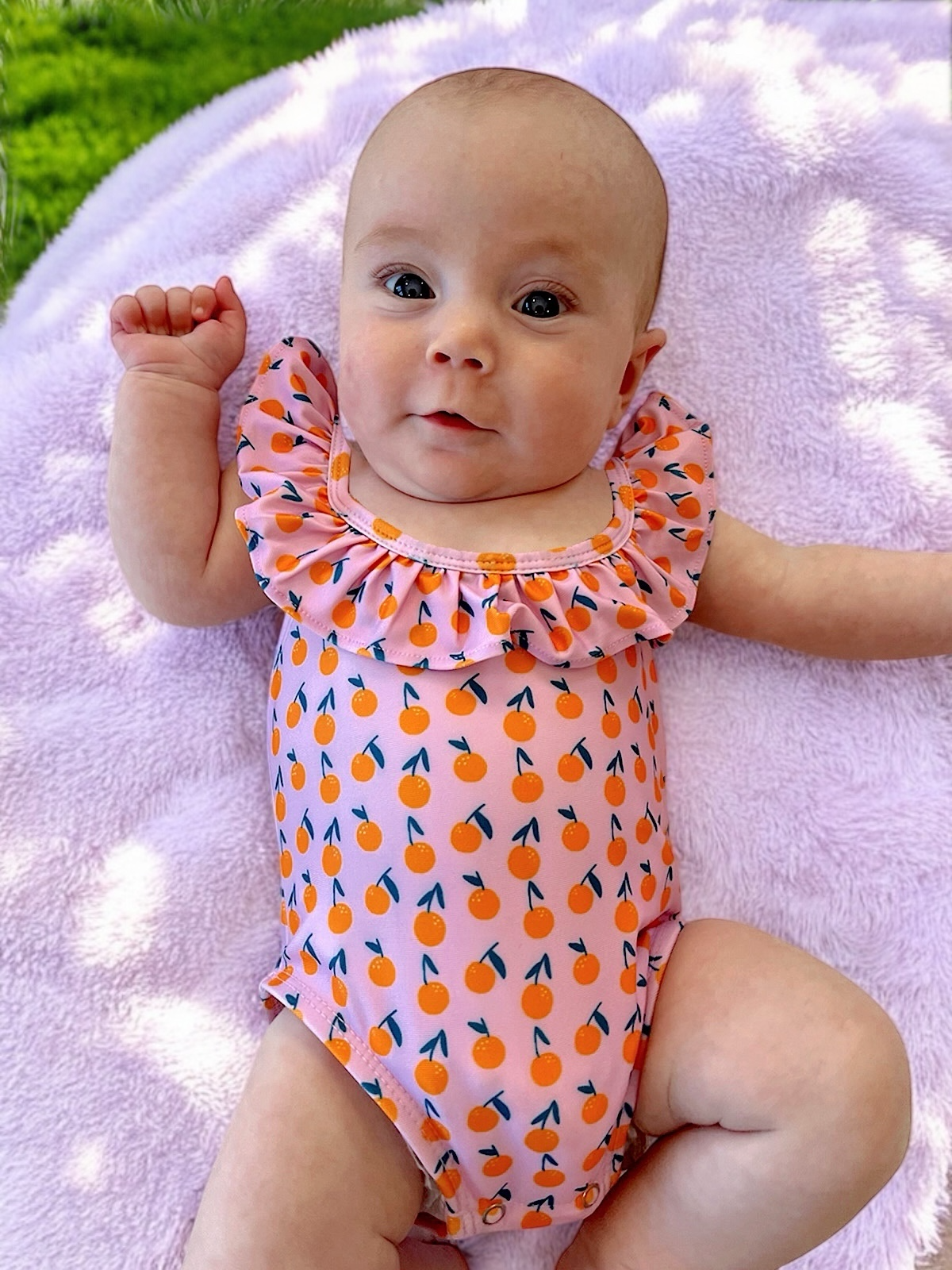Smiling baby in a pink swimsuit with orange patterns, lying on a soft purple blanket outdoors.