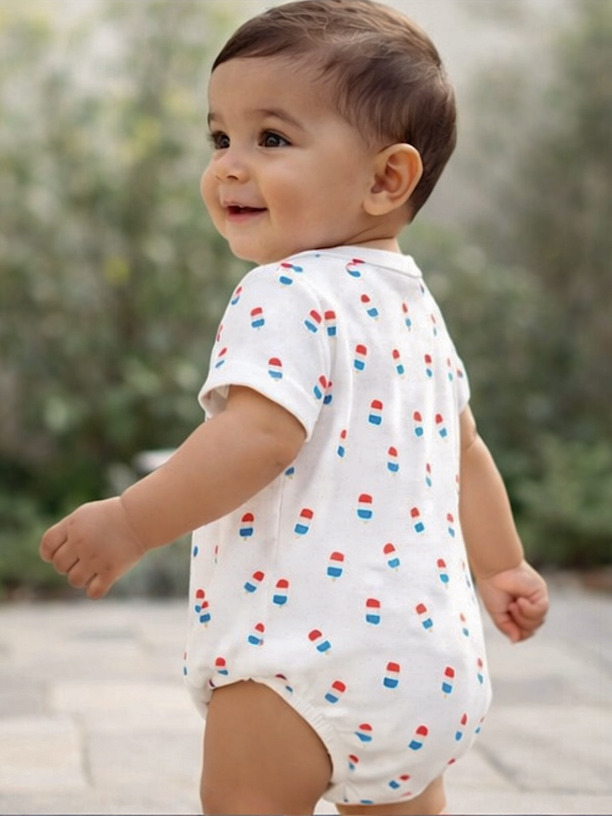 Smiling baby in a patterned onesie, looking back outdoors with greenery in the background.