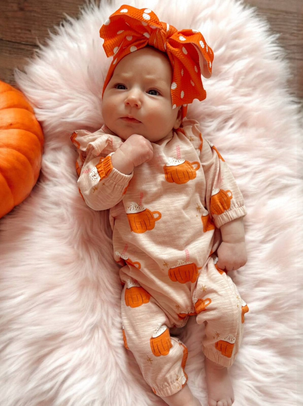 Baby girl in cute pumpkin-themed outfit and large orange polka dot bow, lying on soft pink blanket with a pumpkin nearby.