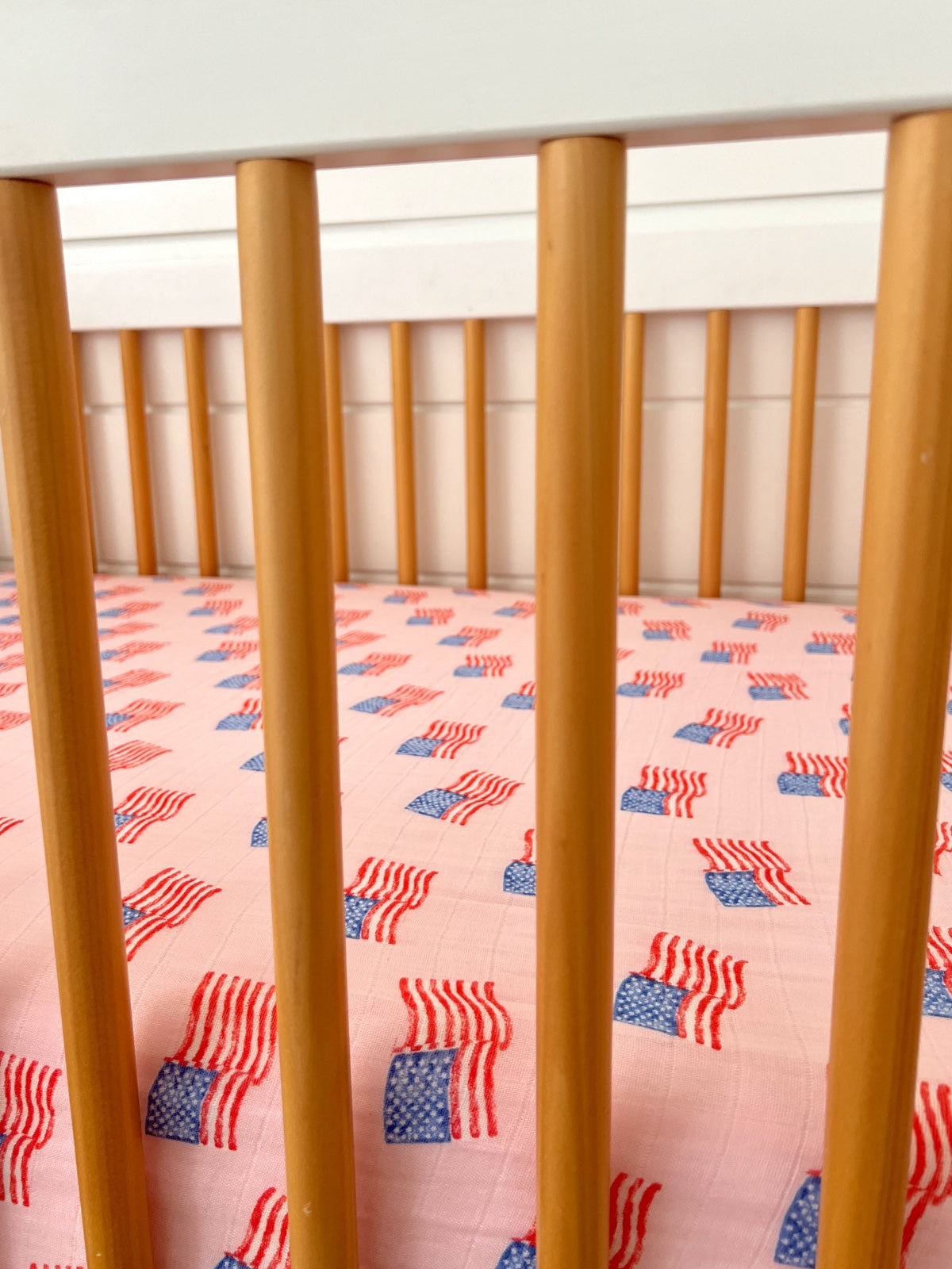 Crib with wooden slats and pink fabric featuring a repeated American flag pattern.