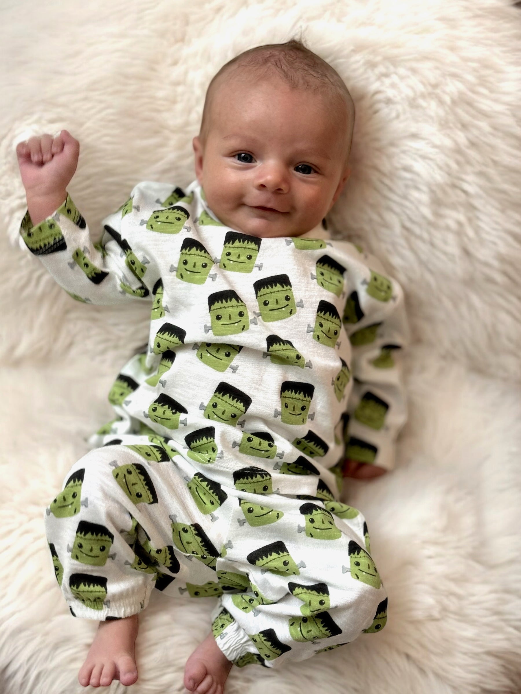 Smiling baby in green Frankenstein-themed pajamas, lying on a fluffy white blanket.