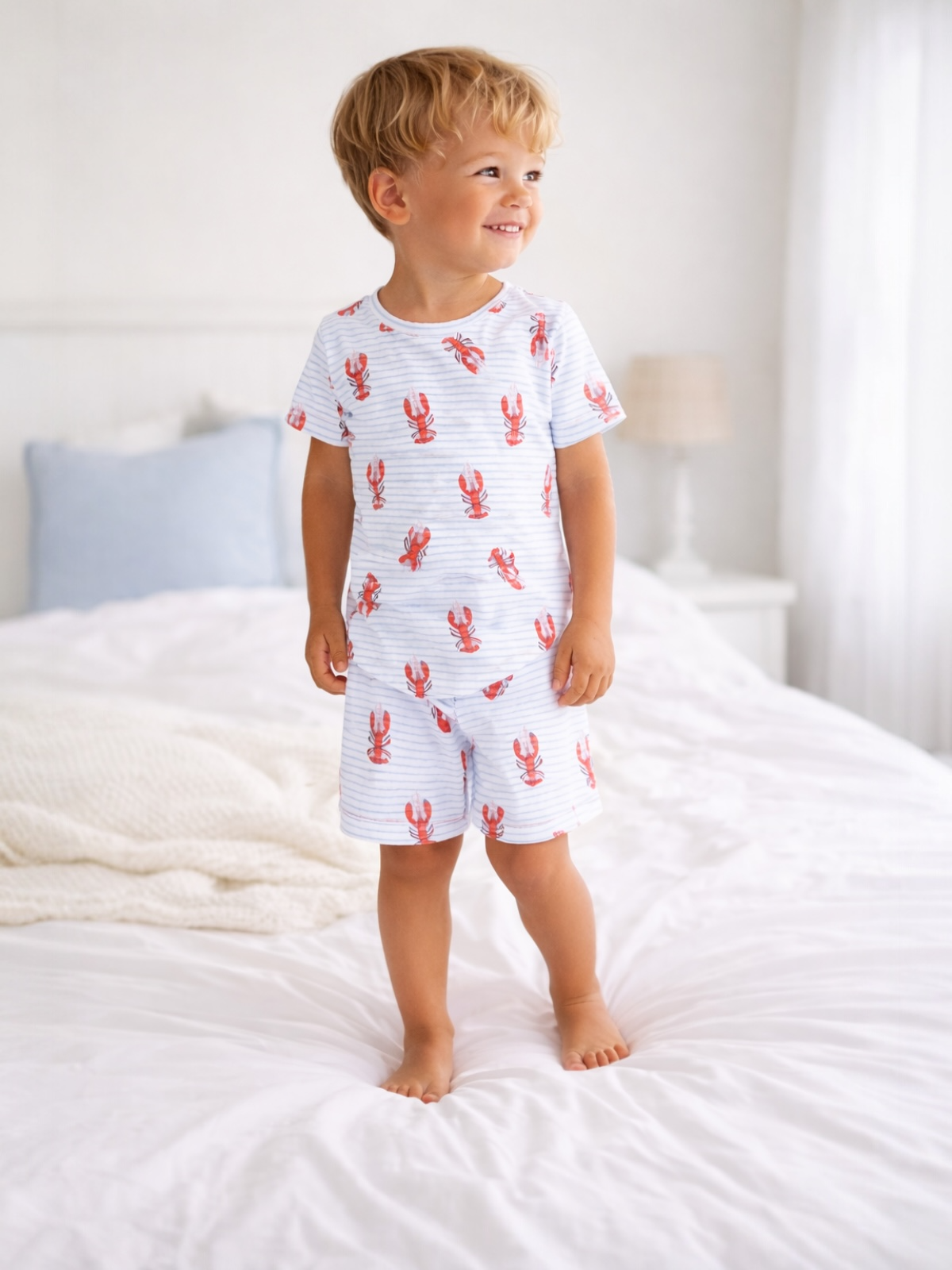 Smiling boy in lobster-patterned pajamas standing on a bed with white bedding and soft natural light.