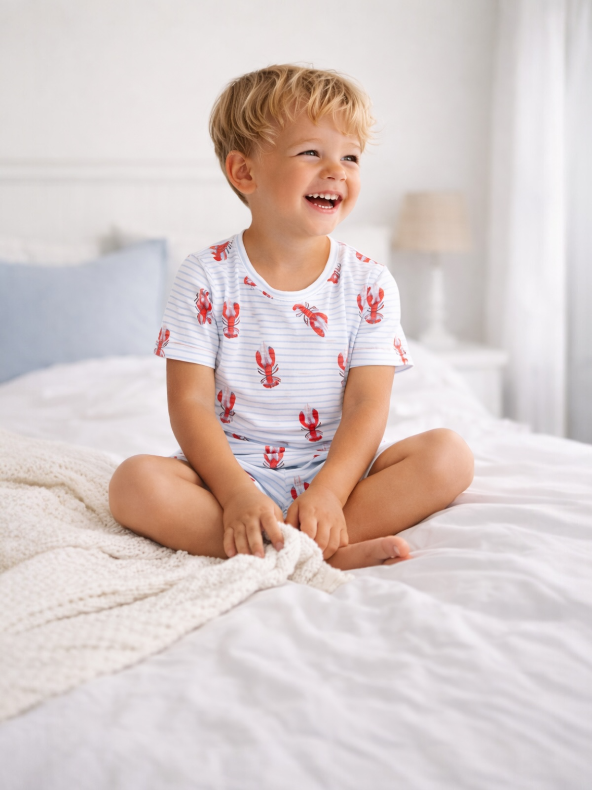 Smiling child in lobster-patterned pajamas sitting on a bed with a cozy blanket. Bright, cheerful atmosphere.