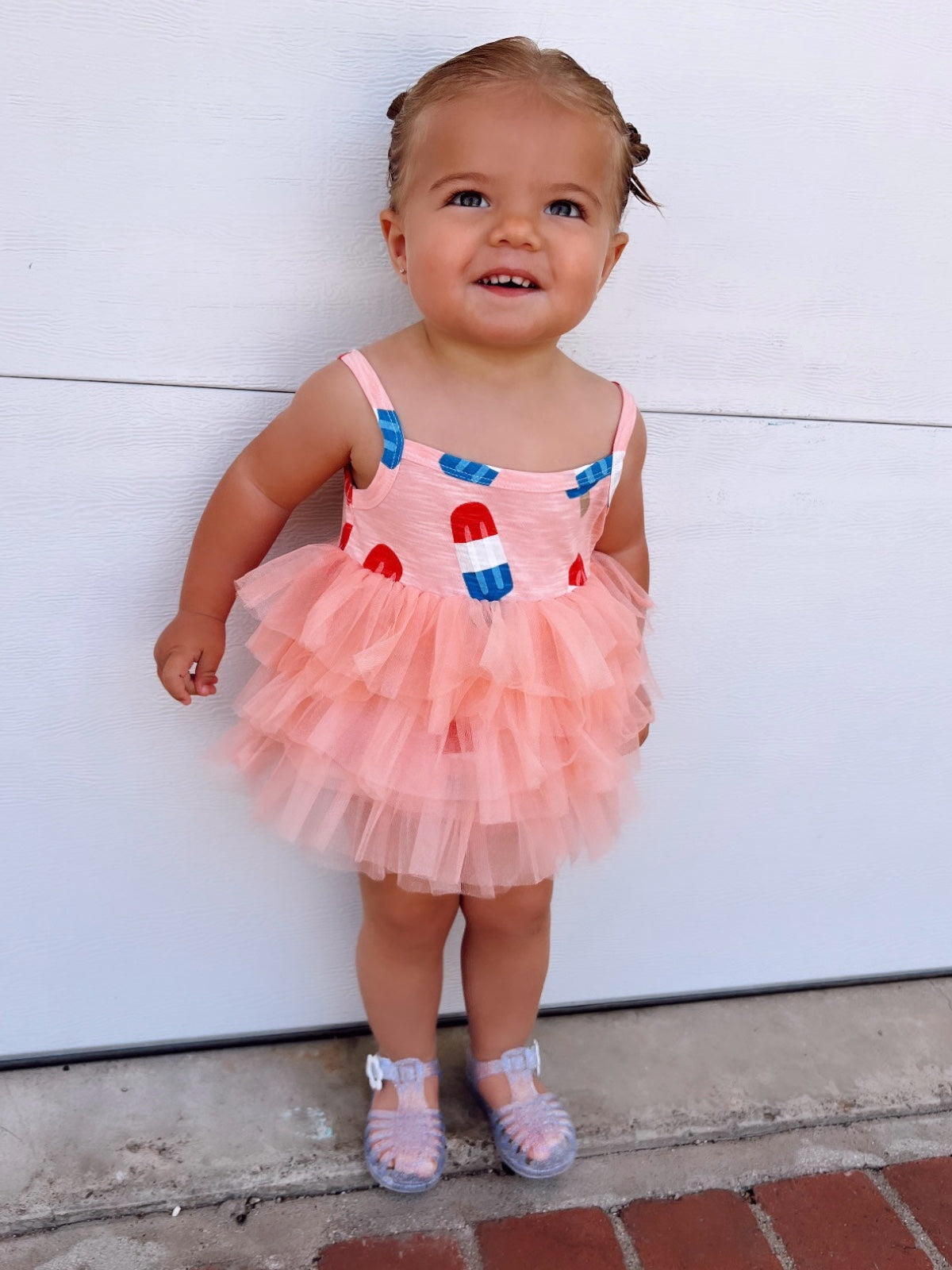Smiling toddler in a pink ruffled dress with popsicle print, standing on a brick walkway near a white wall.