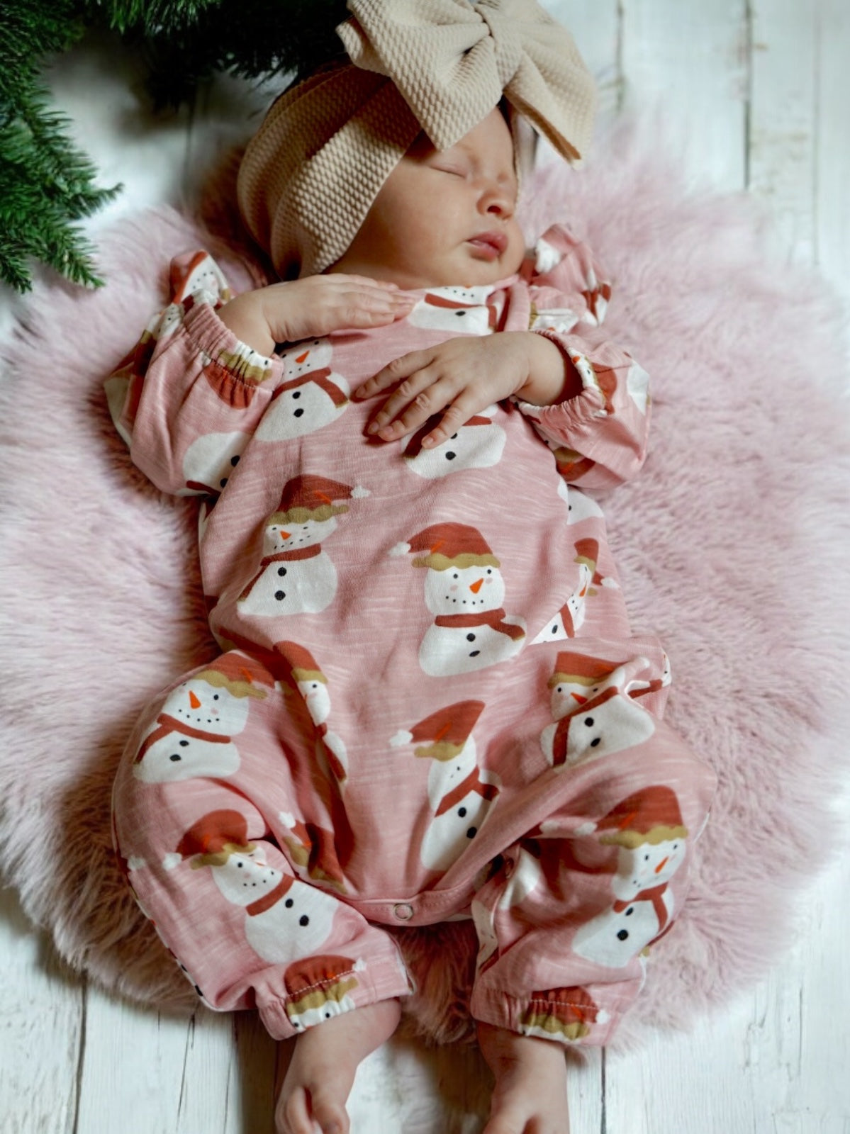 Baby sleeping on a fluffy blanket, wearing a pink snowman onesie and a large beige bow headband.