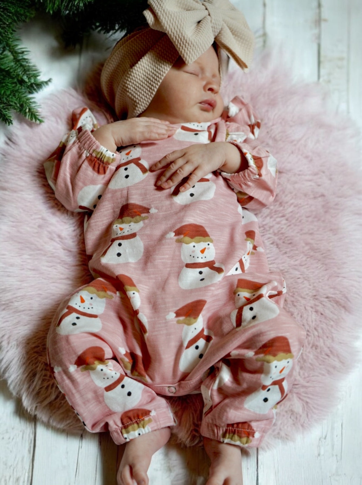 Baby sleeping on a fluffy blanket, wearing a pink snowman onesie and a large beige bow headband.