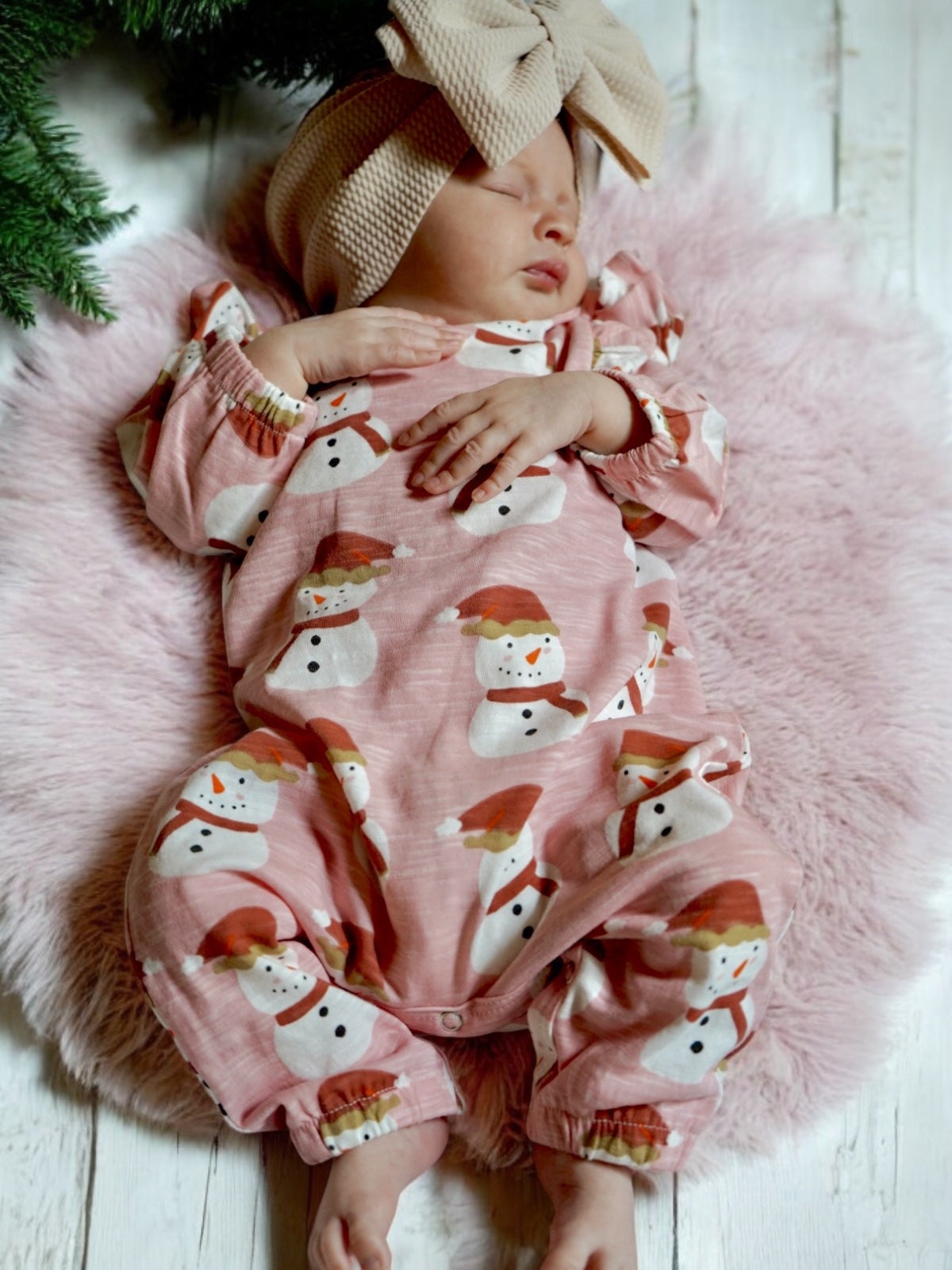Baby sleeping on a fluffy blanket, wearing a pink snowman onesie and a large beige bow headband.