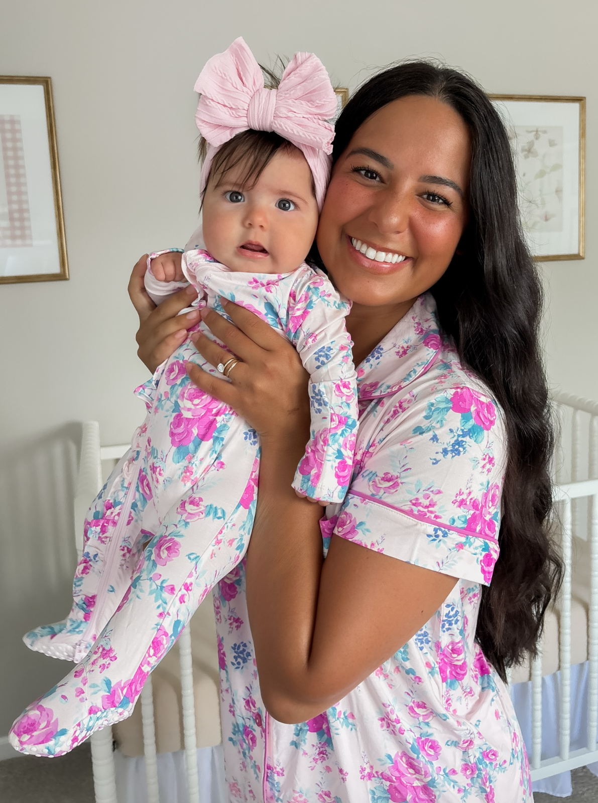 Smiling woman in floral pajamas holds a baby with a large pink bow, against a soft background with a crib.