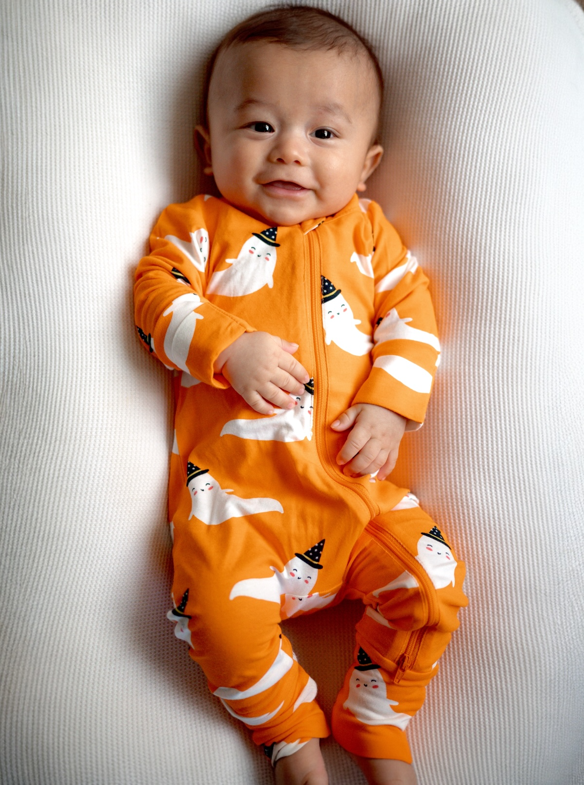 Smiling baby wearing an orange ghost-patterned onesie on a textured white background.