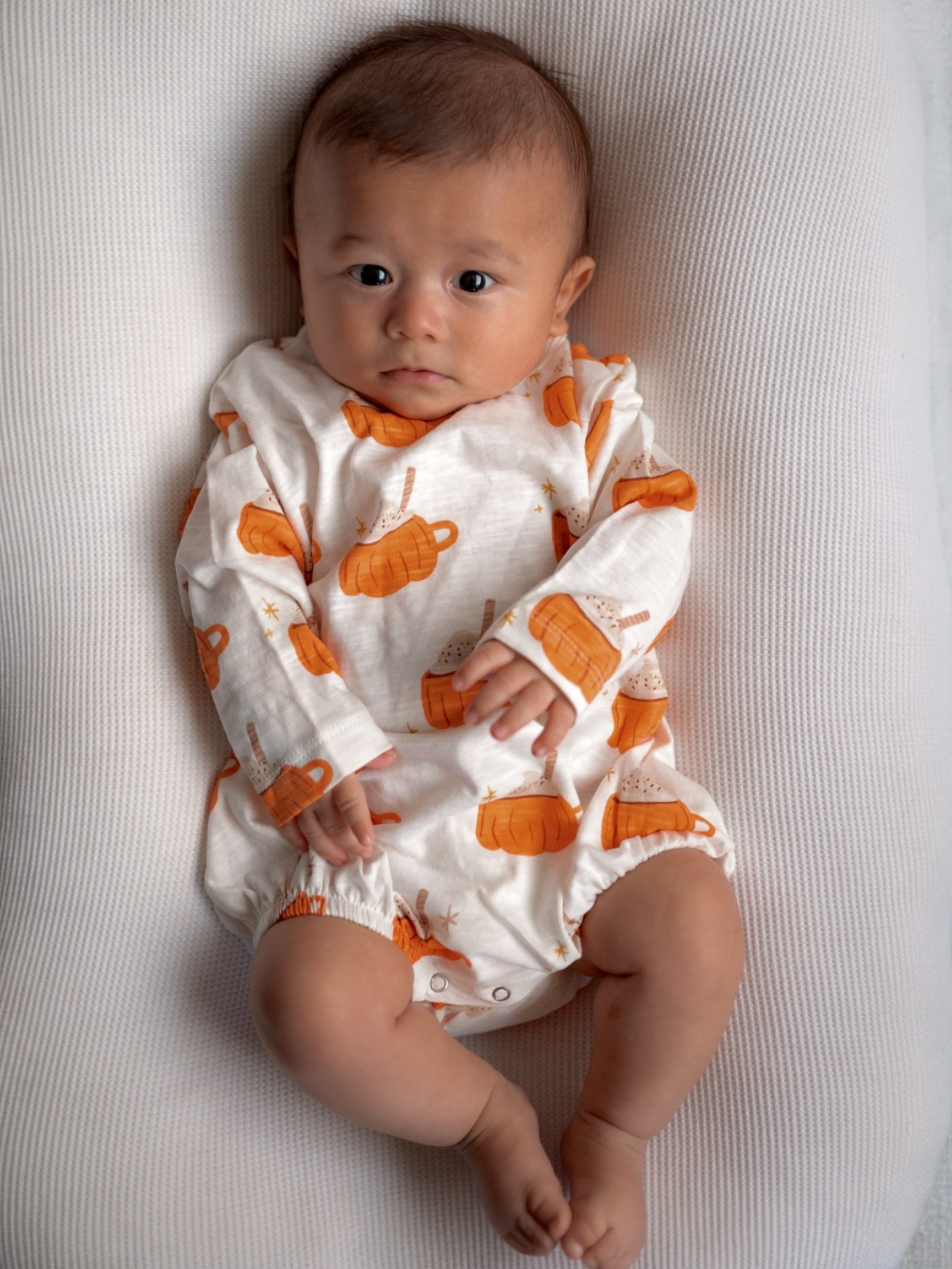 Baby with short hair in a pumpkin-themed outfit, lying on a soft, textured surface, looking directly at the camera.