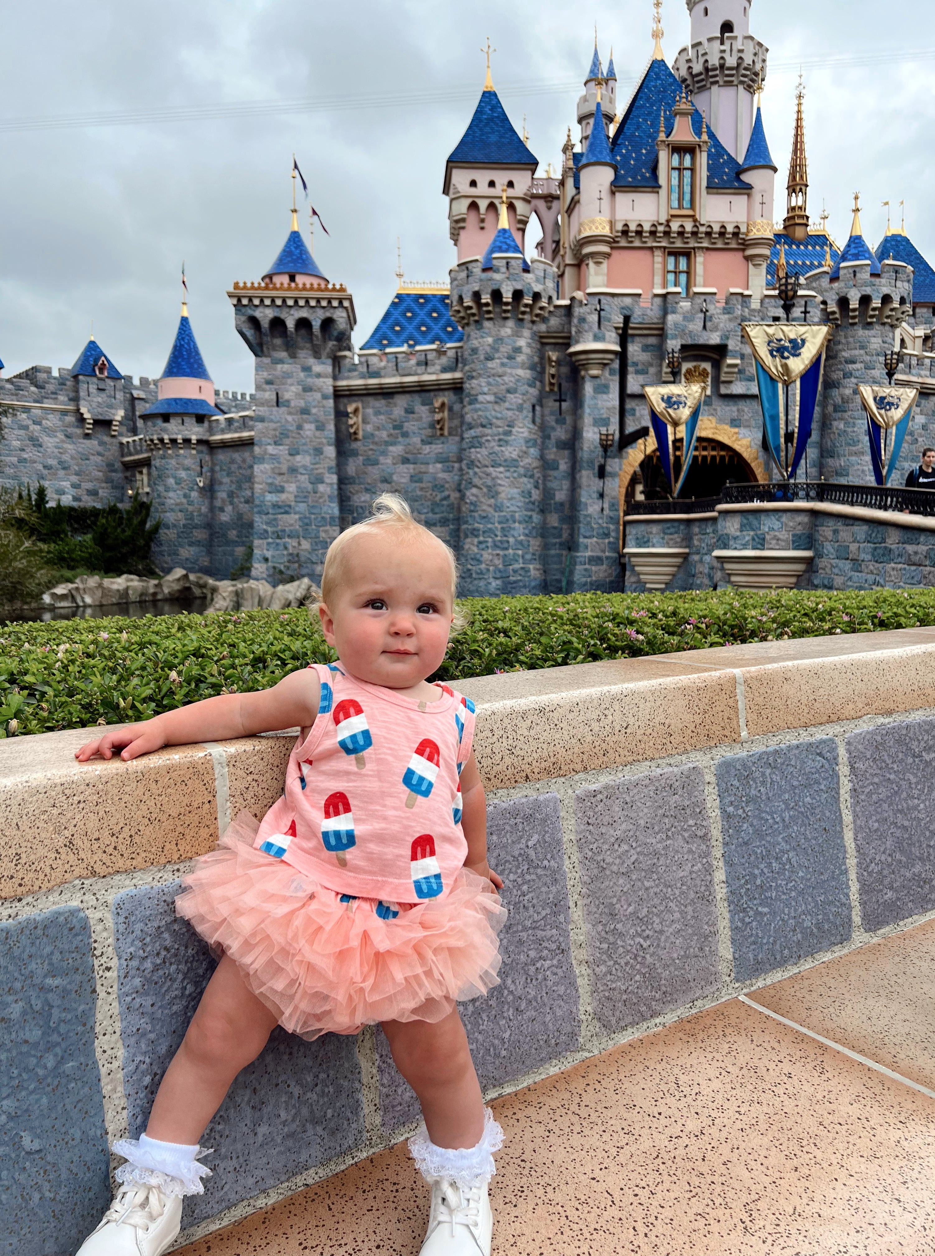 Toddler in a colorful outfit poses in front of a fairytale castle at an amusement park.