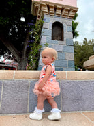 Toddler in a colorful outfit with ruffles, standing by a stone wall in a park setting.