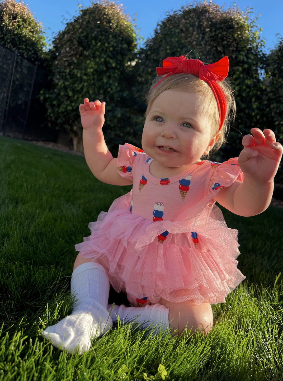 Baby in a pink ice cream-themed dress sits on green grass, smiling with arms raised and a red bow in her hair.