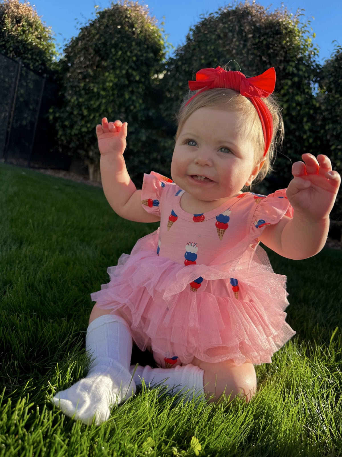 Baby in a pink ice cream-themed dress sits on green grass, smiling with arms raised and a red bow in her hair.