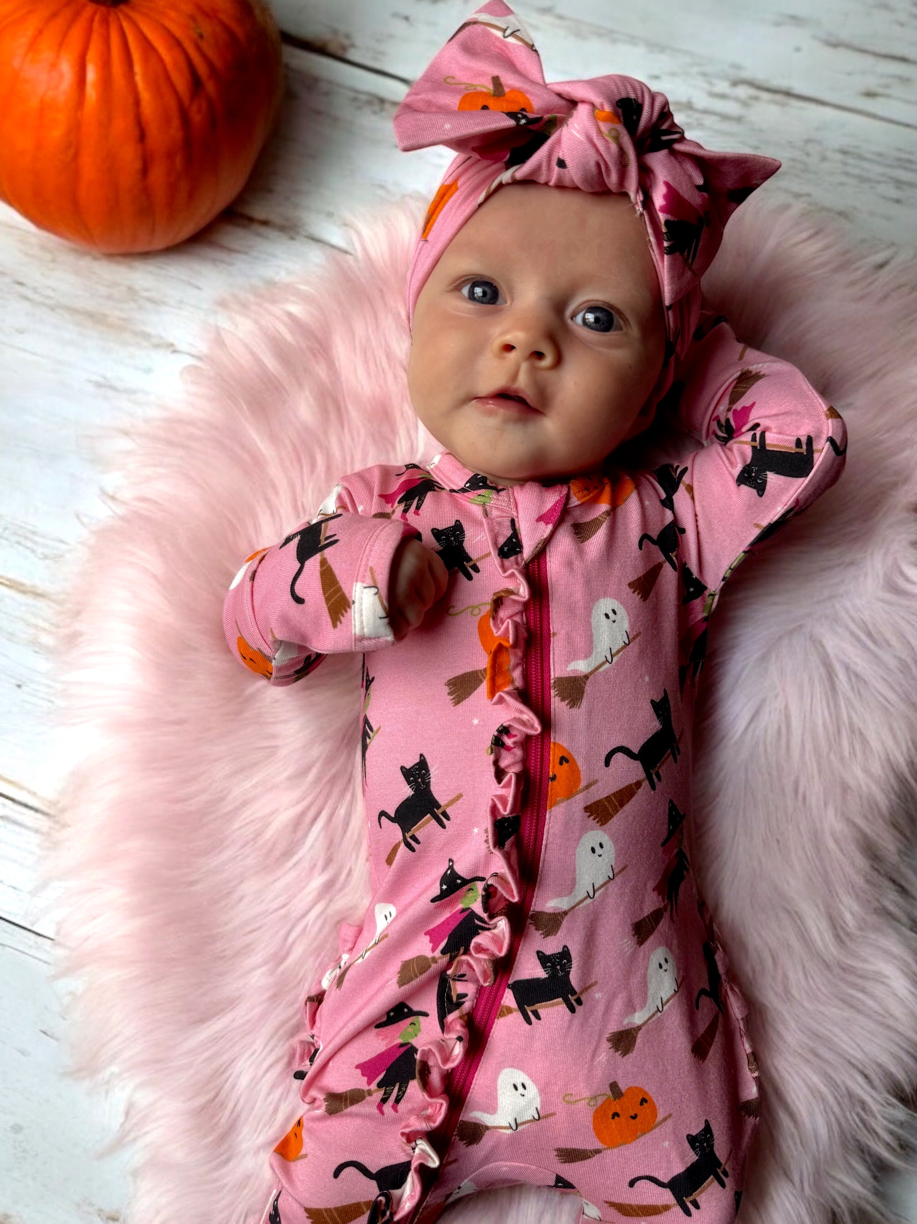 Infant in pink Halloween-themed onesie with cats and pumpkins, lying on fluffy rug beside a pumpkin.