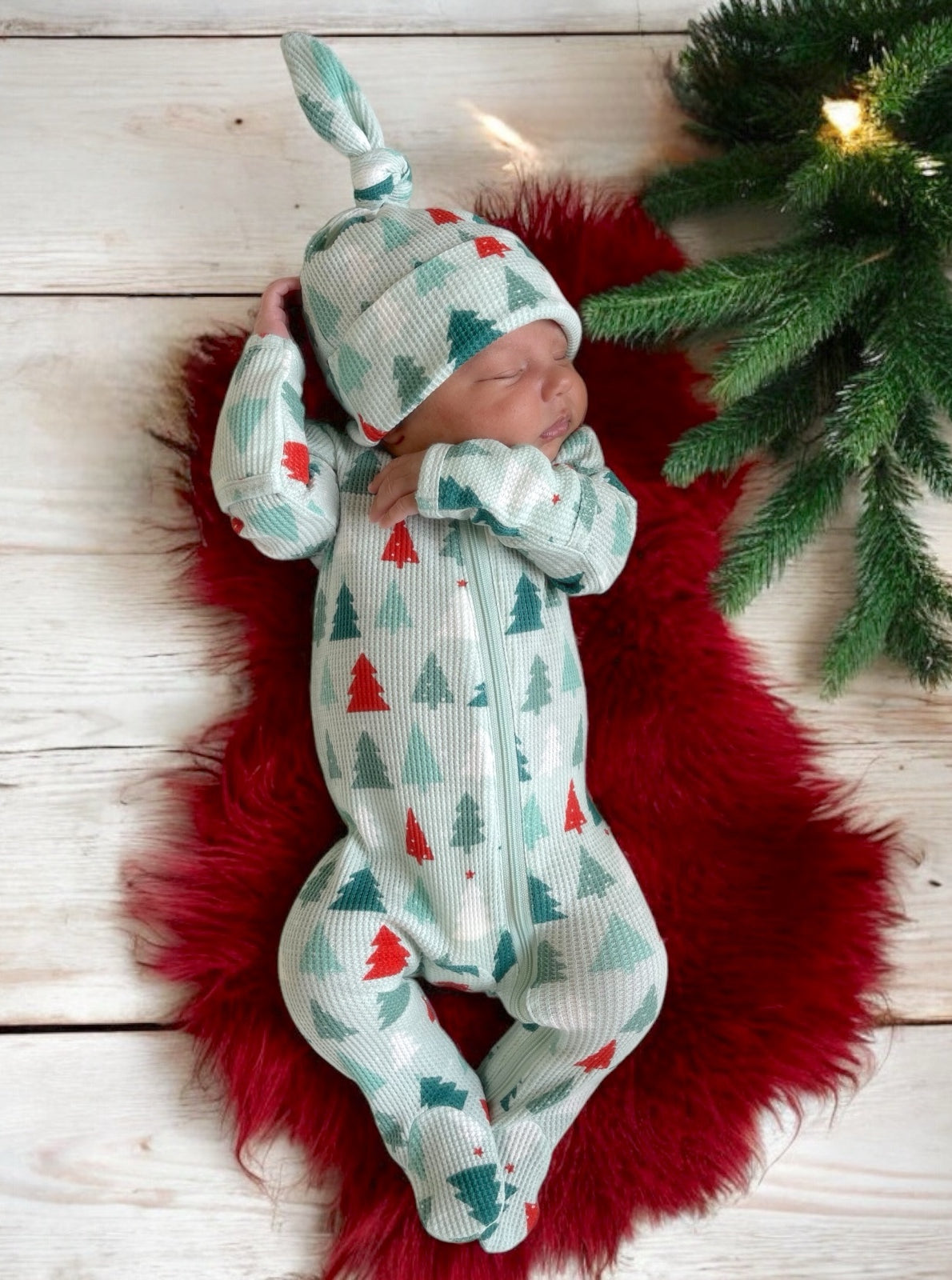 Newborn sleeping on a red rug, wearing a mint green outfit with red and teal trees, and a matching hat.