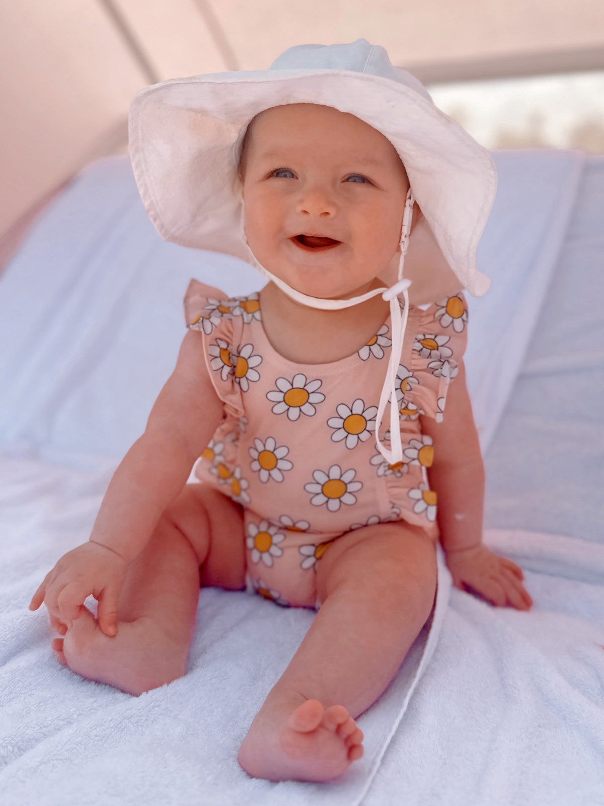 Smiling baby in a floral swimsuit and sun hat, sitting on a towel in a sunlit setting.