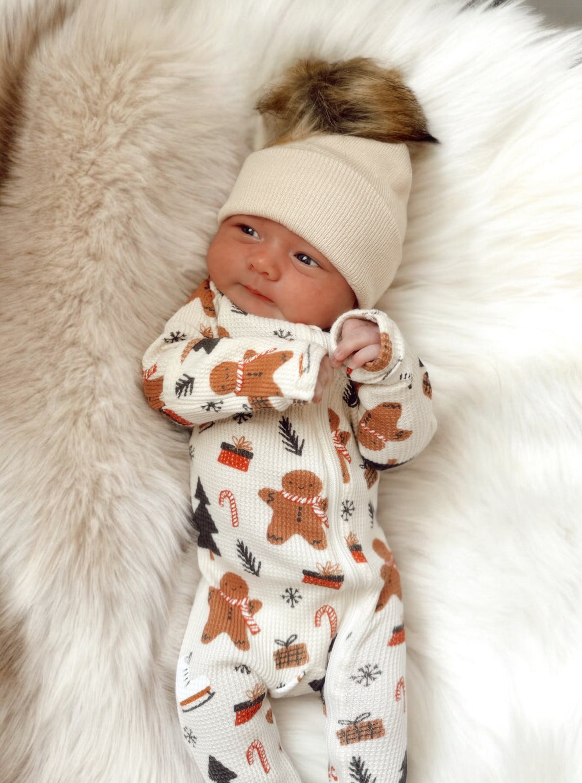Newborn in cozy gingerbread-themed outfit, wearing a beige hat with a fur pompom, resting on fluffy white fur.